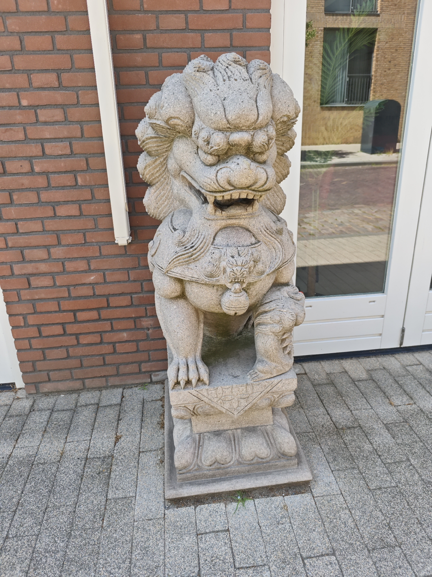 The image shows a stone statue of a Foo dog, or guardian lion, standing on a cobblestone pavement in front of a building. The Foo dog is grey, intricately carved, and appears to be weathered. It has a fierce expression with its mouth open, revealing its teeth. The building behind it has a brick facade on the left and a large glass door on the right, reflecting the surroundings. The pavement is made of rectangular grey stones. The scene is outdoors, and the lighting suggests it is daytime. This statue is located in Tilburg, Netherlands.