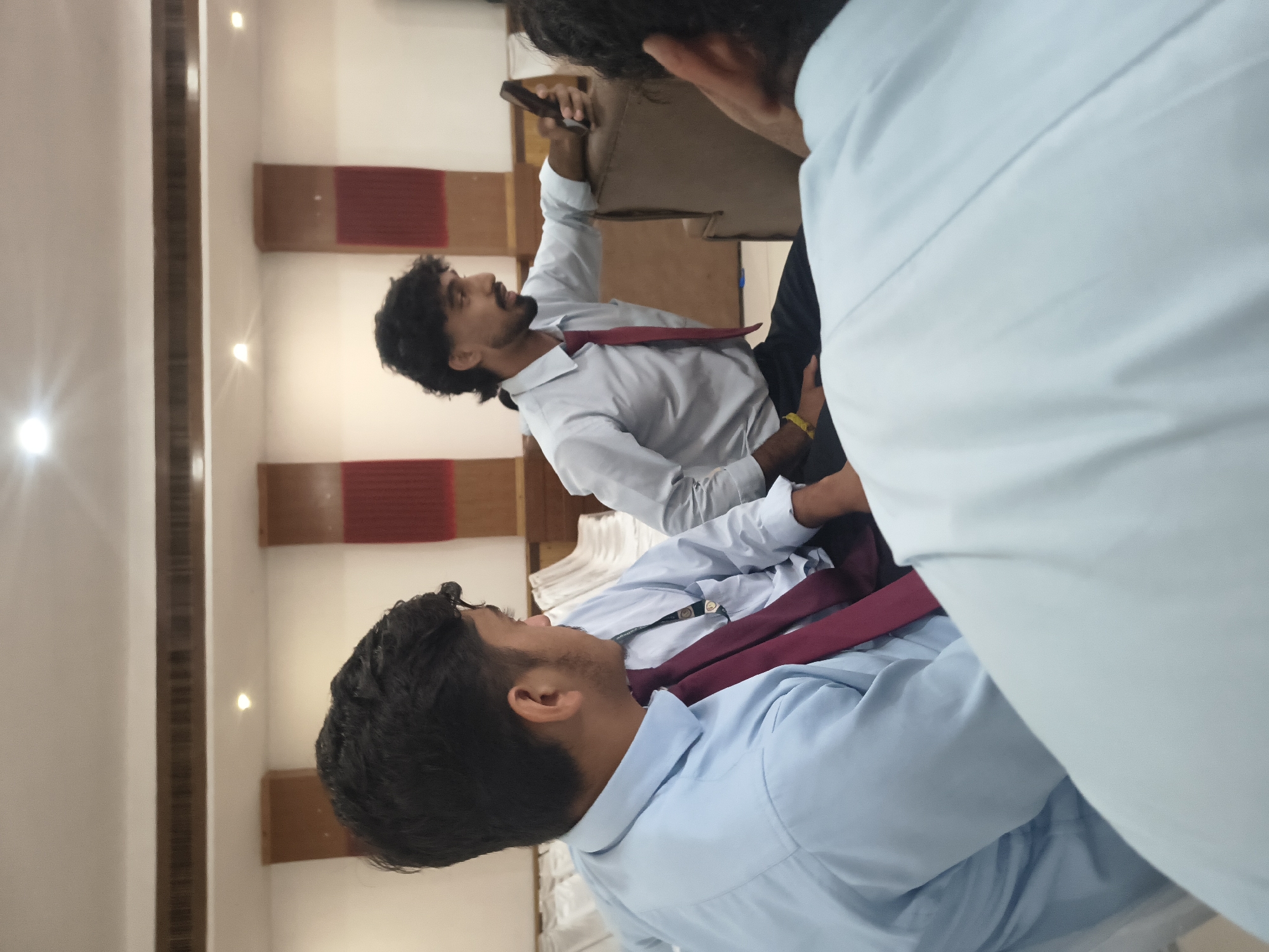 In Khaniara, India, three young men are seated indoors, appearing to be in a hall or meeting room. The man in the foreground, facing left and out of focus, wears a light blue collared shirt with a dark red tie. To his right, another man, also wearing a light blue shirt and a dark red tie, is in sharper focus, looking at a dark-colored mobile phone he is holding in his right hand. He has a beard and dark, wavy hair. Behind these two men, a third individual, mostly out of frame, is visible, also wearing a light blue collared shirt. The background features white walls with decorative panels, warm-toned wooden accents on the ceiling, and what appear to be rows of chairs draped in white fabric. The lighting suggests an indoor event, possibly a formal gathering or business meeting.