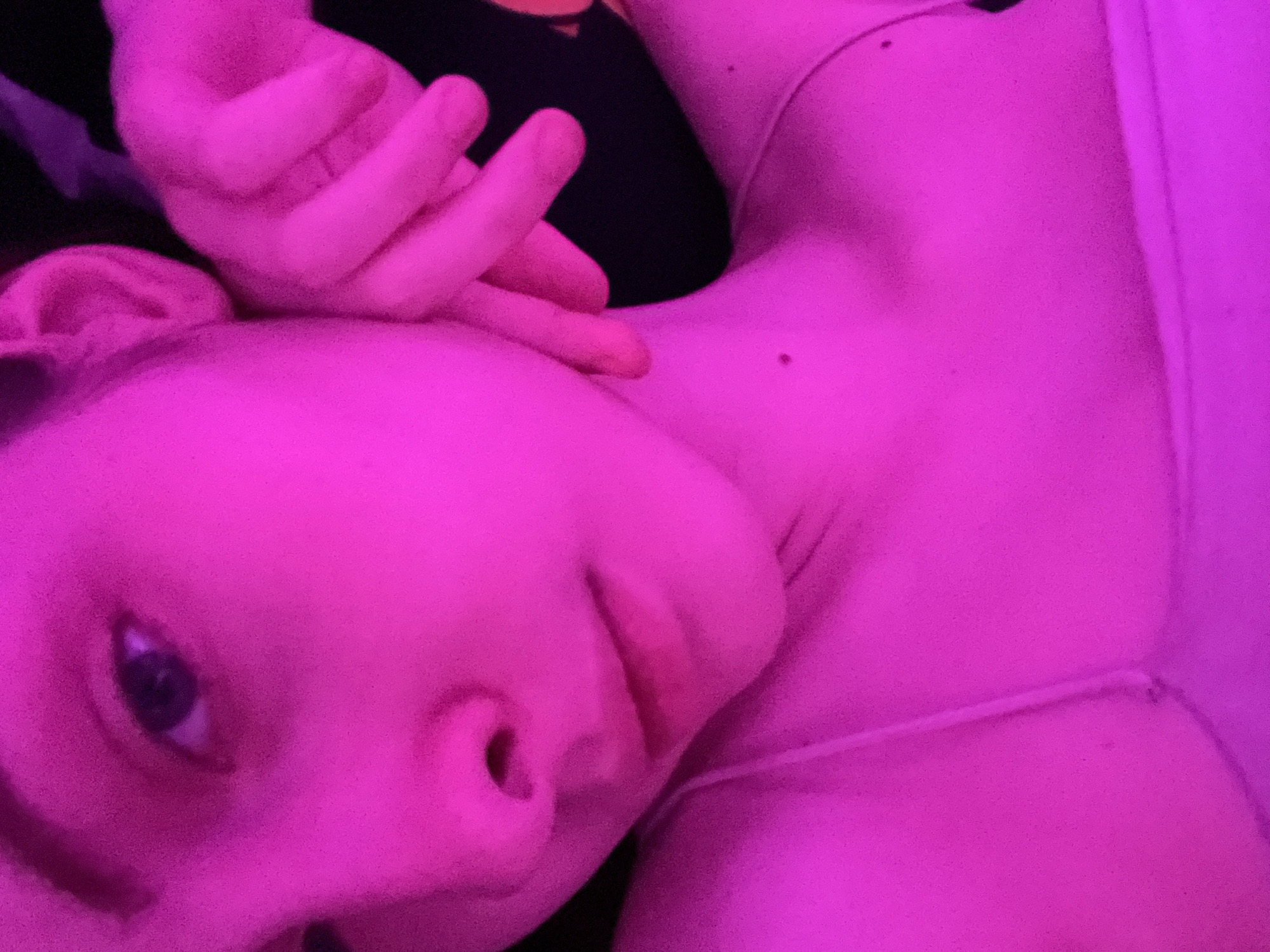 The image shows a person lying down, with their face and upper chest visible. The person's head is tilted to the side, and they have one hand near their face. They are wearing a light-colored top with thin straps. The lighting in the image is pink, affecting the overall color cast. The background is dark and mostly obscured.
There is no indication of the location being Saint Petersburg, Russia or any city location.