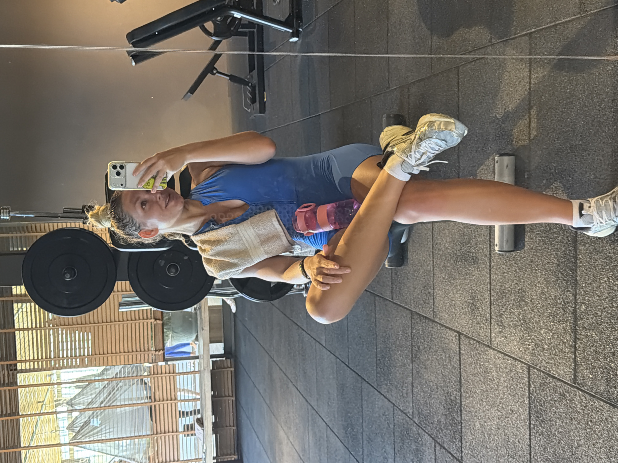 The image depicts a woman taking a selfie in what appears to be a gym. She is wearing a blue athletic outfit and white sneakers. A towel is draped over her shoulder, and she is holding a water bottle. Weightlifting equipment, including a barbell with weights and a bench, is visible in the background. The gym has a grey tiled floor and a partially obscured view of the outside, showing a structure with wooden slats and a glimpse of a net. There is no definitive information to conclusively place this image in Alanya, Turkey. The location details are absent in the image.