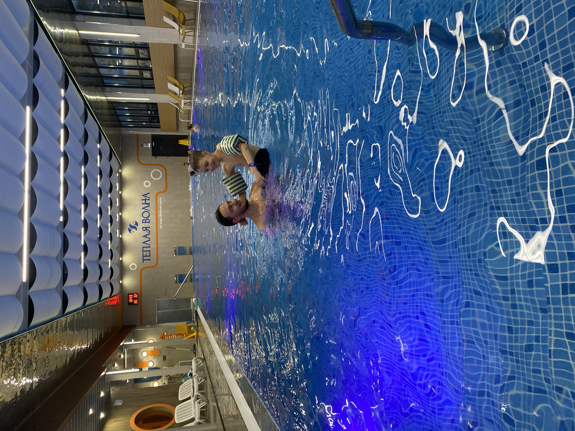 A medium shot shows a man and a child in a swimming pool. The man, with dark hair and a beard, is in the water up to his chest. The child, wearing a striped green and white top and a dark bottom, is being held by the man, appearing to be supported on the man's shoulders. The pool has blue tiles and white markings that appear to be decorative. In the background, out of focus, are other figures in the water, pool-side furniture, walls, lights, and a sign with Russian text, "ТЕПЛАЯ ВОЛНА" (TEPLAYA VOLNA). This photo was most likely taken in the city of Votkinsk, Russia.