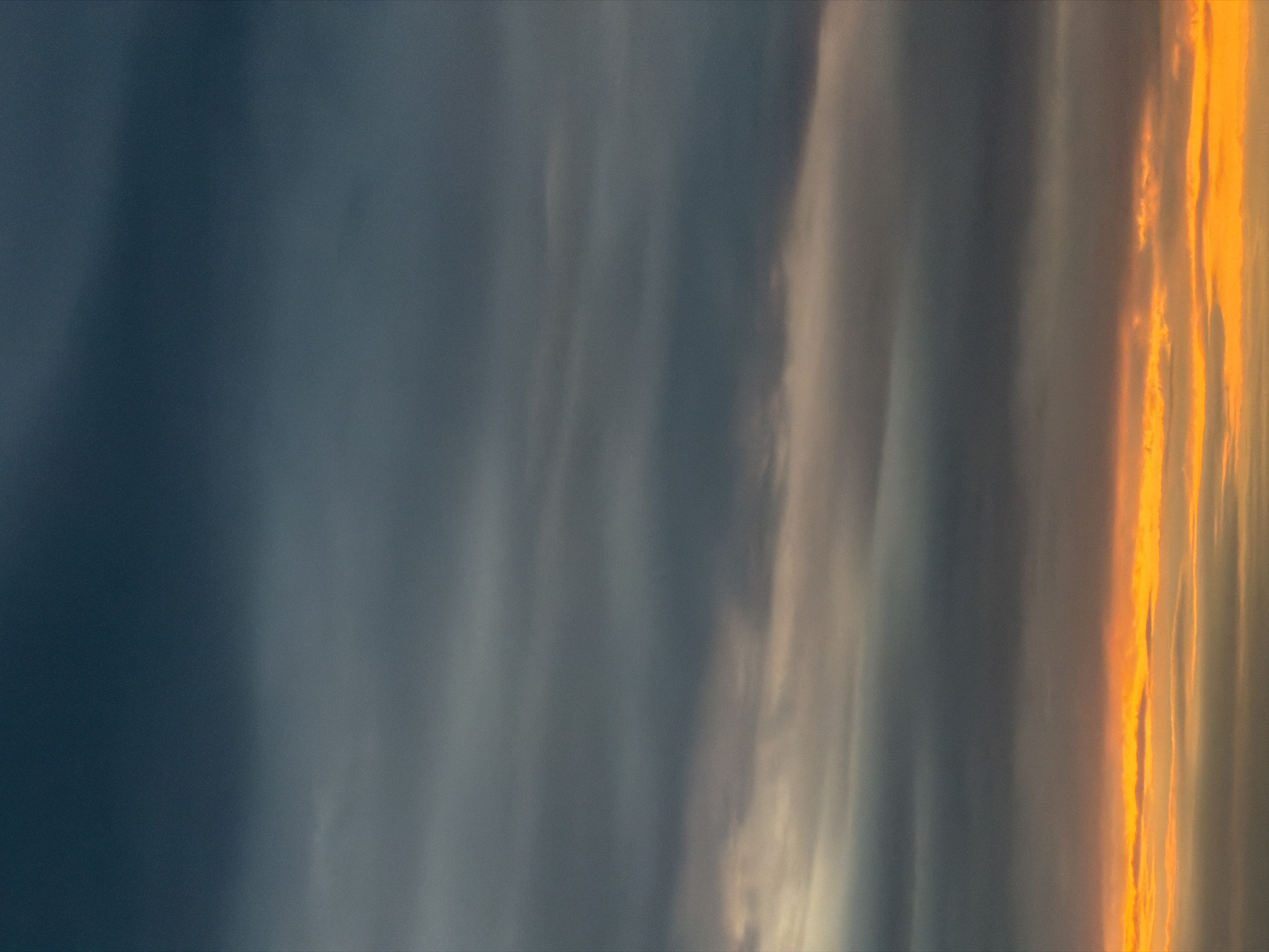 The image depicts a sky, possibly at dawn or dusk, with a significant portion dominated by dark, striated clouds. To the right, the clouds are illuminated with a bright orange color, suggesting the presence of the sun either rising or setting. The image's orientation is vertical, as if the camera was pointed upwards. There are no clear indications of a city or people in the image.