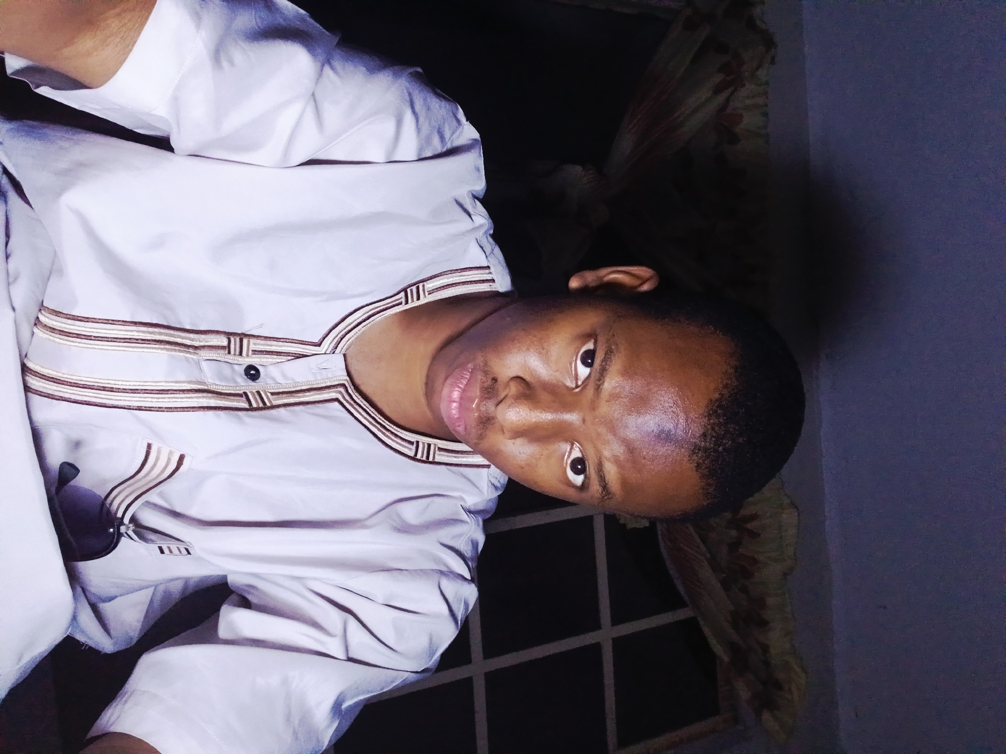 A young man of African descent is captured in a close-up, waist-up selfie. He is wearing a light-colored, short-sleeved traditional outfit adorned with brown and white striped embroidery around the neckline and down the front. A pair of dark sunglasses is tucked into the chest pocket of his garment. The background is dimly lit, featuring a patterned curtain and a window with a grid-like structure. The overall lighting suggests an indoor setting, possibly at night, with the light source appearing to be from the front, casting a slight shadow on the wall behind him. The man's facial expression is neutral. The setting is consistent with a residential environment within the city of Lagos, Nigeria.