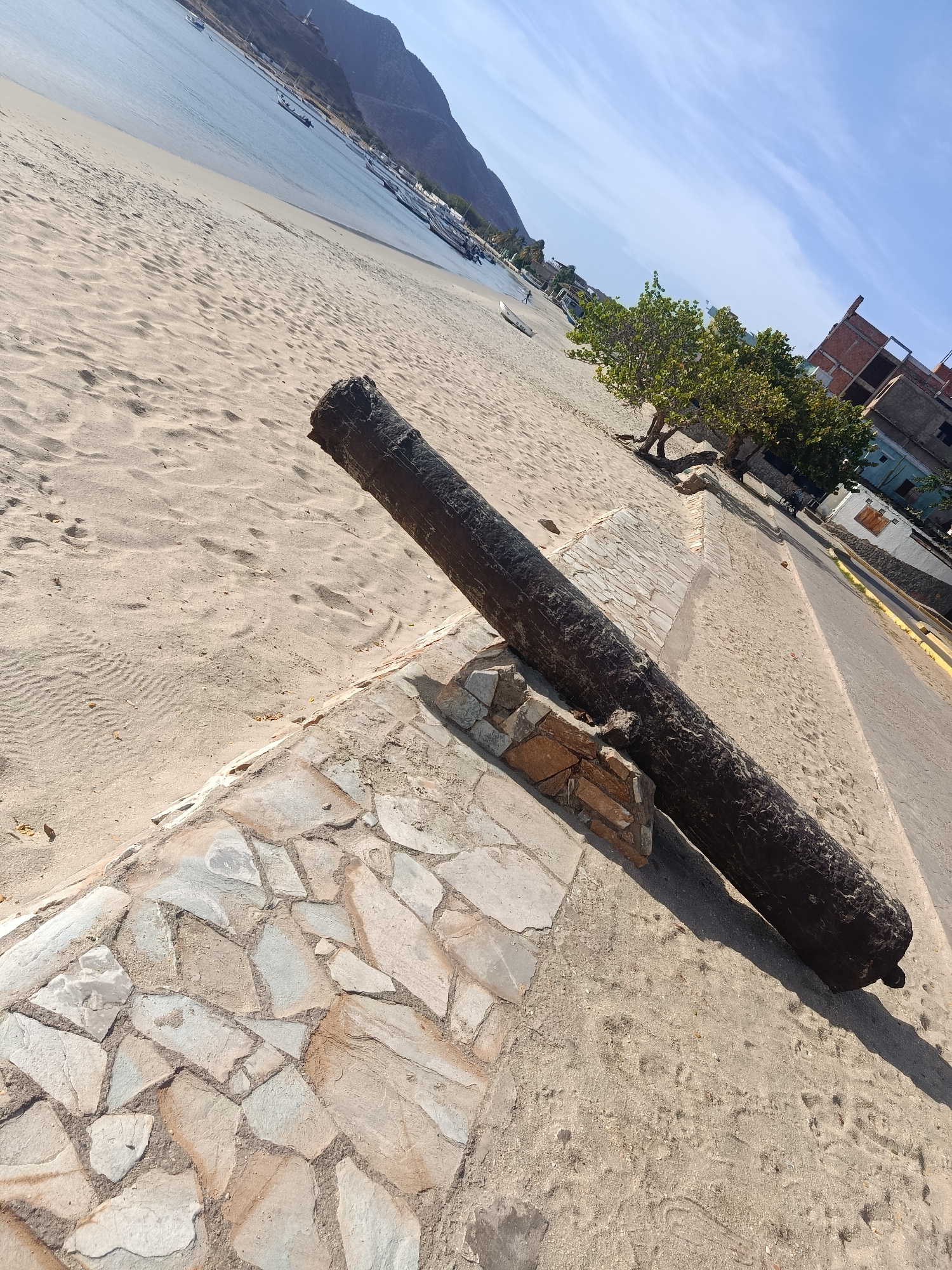 The image captures a scene in Juan Griego, Venezuela, featuring a historical cannon prominently positioned near the shoreline. The cannon rests on a stone base, situated between a sandy beach and a paved walkway. The beach stretches towards the water, where several boats are visible, set against a backdrop of mountains. Adjacent to the walkway, some trees and buildings are present, suggesting an urban area close to the beach. The sky is clear and blue, indicating a sunny day. No people are immediately visible in the frame.