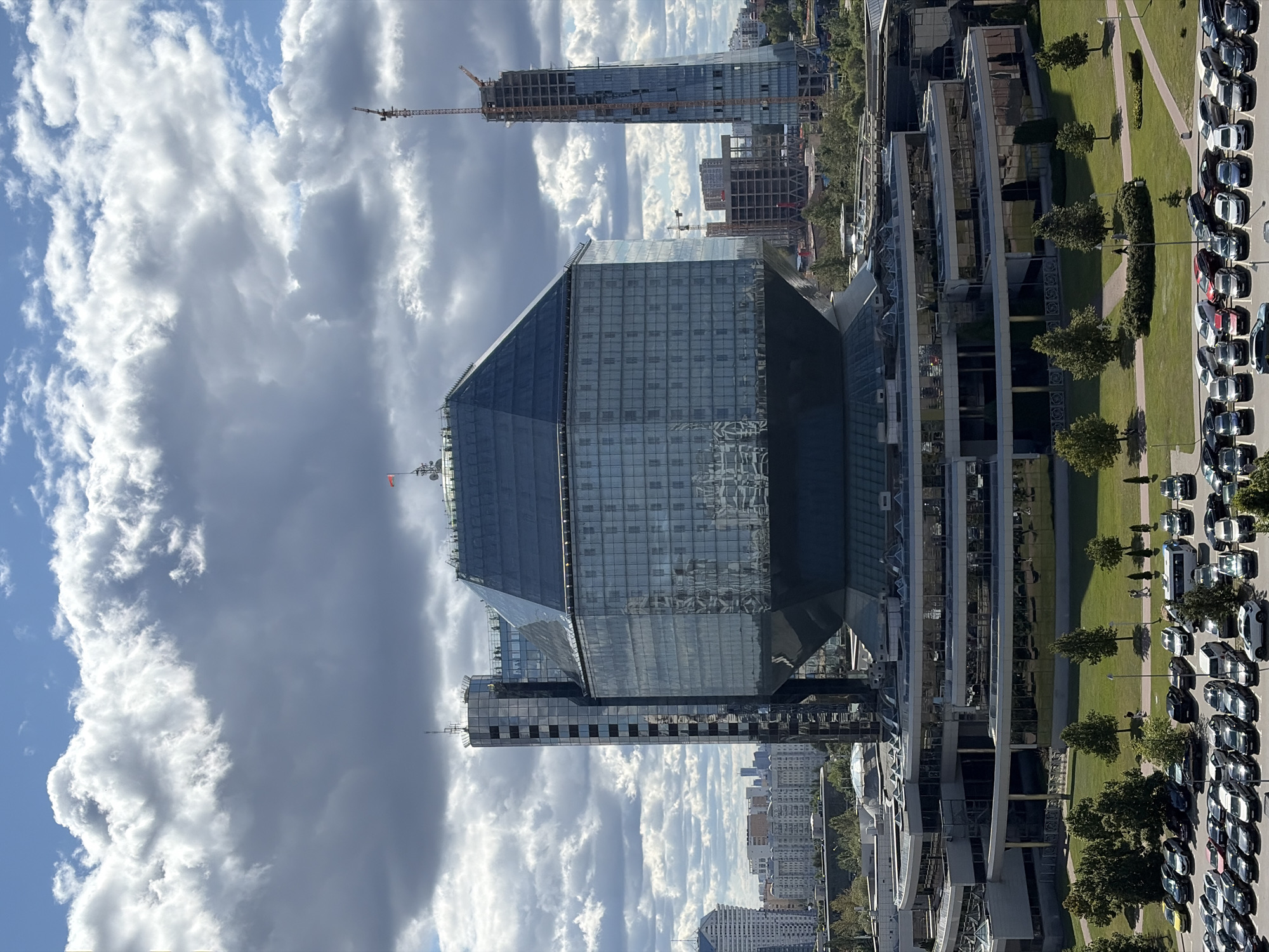The image shows a wide shot of the National Library of Belarus, a distinctive faceted building with a glass exterior, located in Minsk, Belarus. The library is situated in a suburban area with other buildings and construction sites visible in the background. The foreground shows a parking lot filled with various cars, and a well-maintained green lawn with scattered trees and walking paths. Two people are walking on one of the paths. The sky is partly cloudy with a mix of white and gray clouds. The overall scene depicts a modern urban landscape with elements of green space and ongoing development.