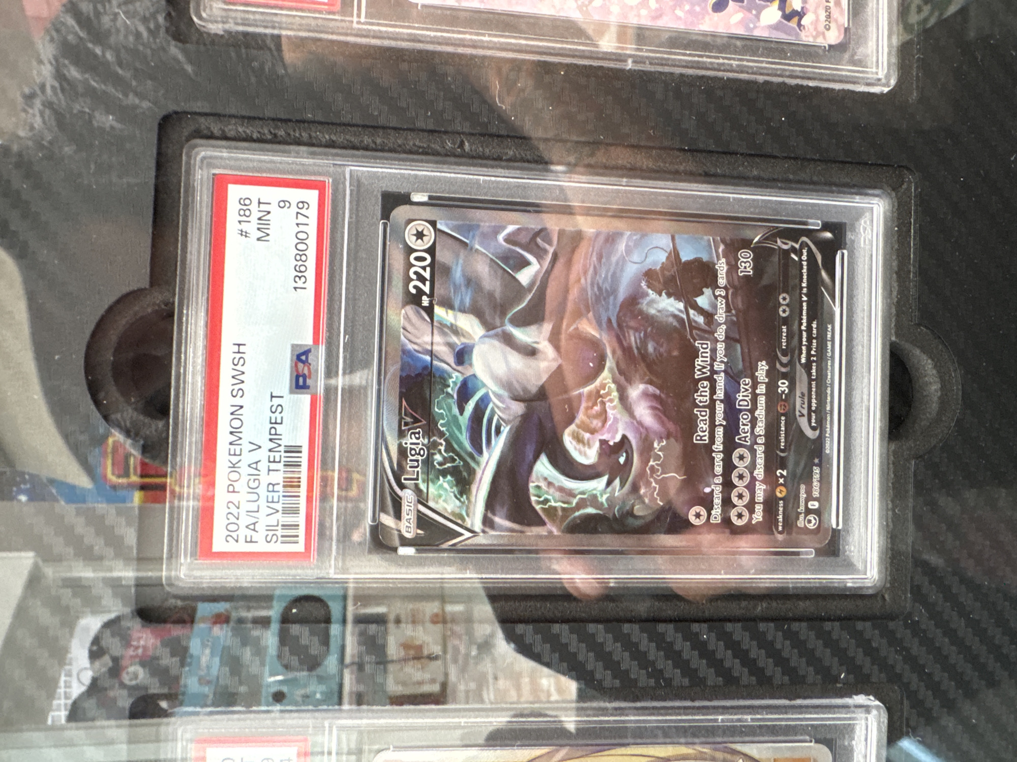 A PSA-graded Pokemon card of Lugia V from the Silver Tempest set is displayed. The card is in a clear protective case, with a red and white PSA label on top. The label indicates the card is #186, MINT, graded a 9, with the serial number 136800179. The card itself features an illustration of Lugia with stormy effects. The surrounding environment is dark, with some reflected light suggesting an indoor display case. The location of El Paso, United States of America, is not discernible from the image.