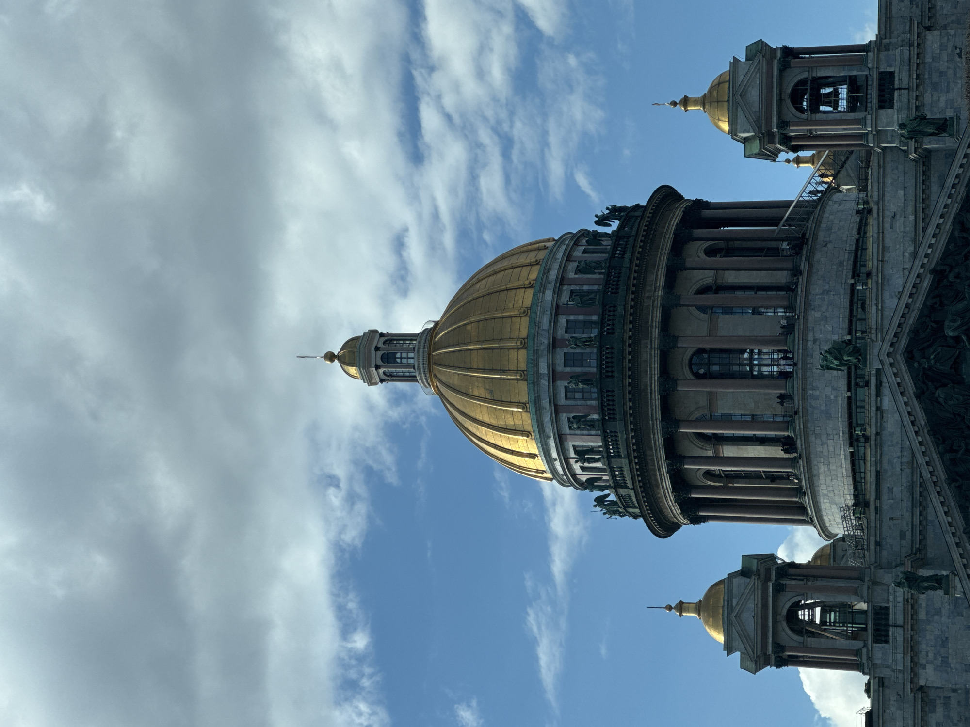 The image depicts a portion of Saint Isaac's Cathedral in Saint Petersburg, Russia. The golden dome is prominent, contrasted against a cloudy sky. Part of the cathedral's exterior, including architectural details like columns and statues, is visible. Two smaller domes atop towers are visible on either side of the central dome. The photograph is taken from a low angle, emphasizing the height and grandeur of the cathedral.