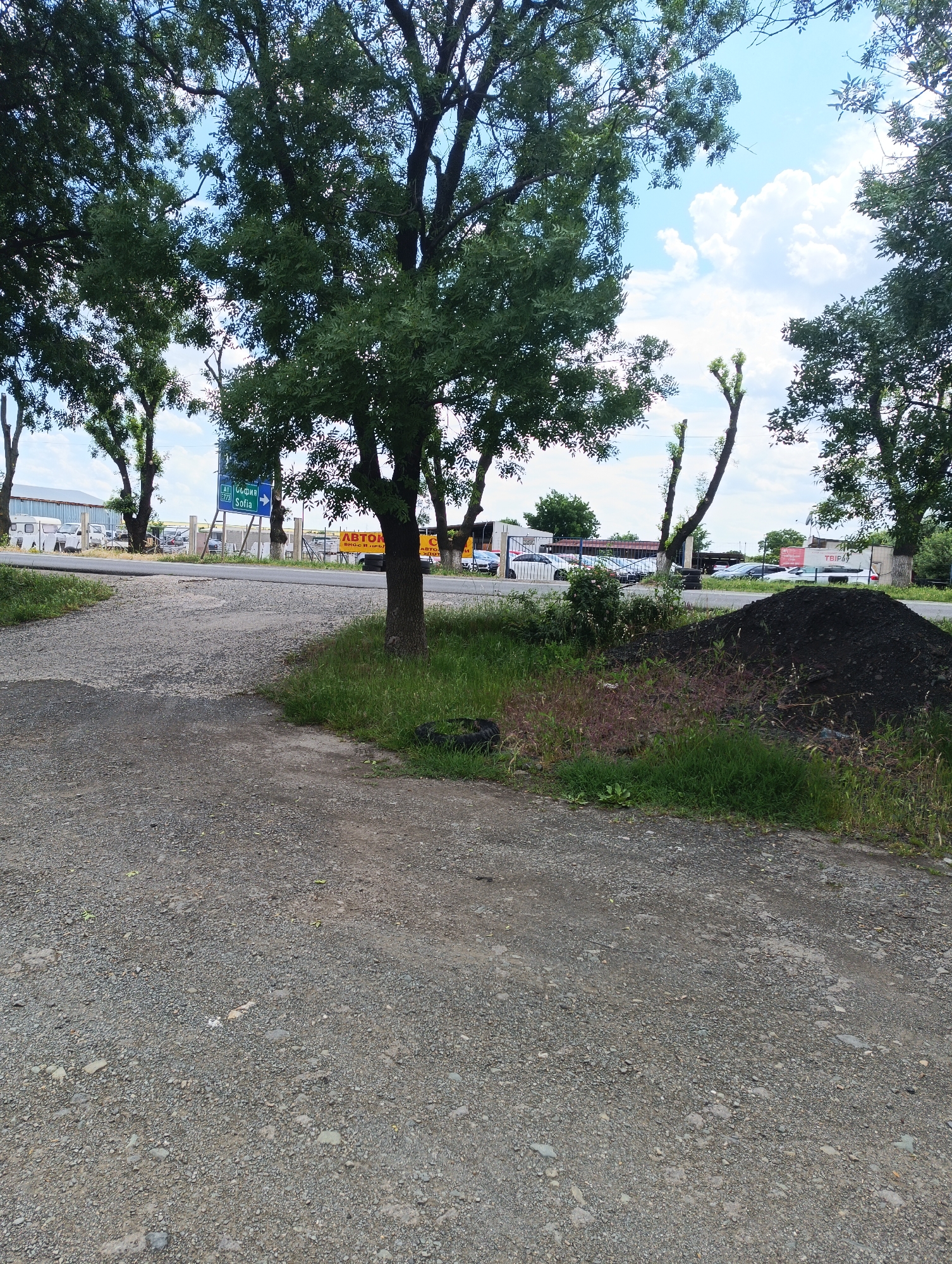 The image shows a gravel road in Burgas, Bulgaria, leading to a T-intersection with a paved road. There are trees along the roadside. A blue and green sign indicates the direction to Sofia. A pile of dirt is on the right side of the road, near a cut-down tree. Cars are parked in the background.