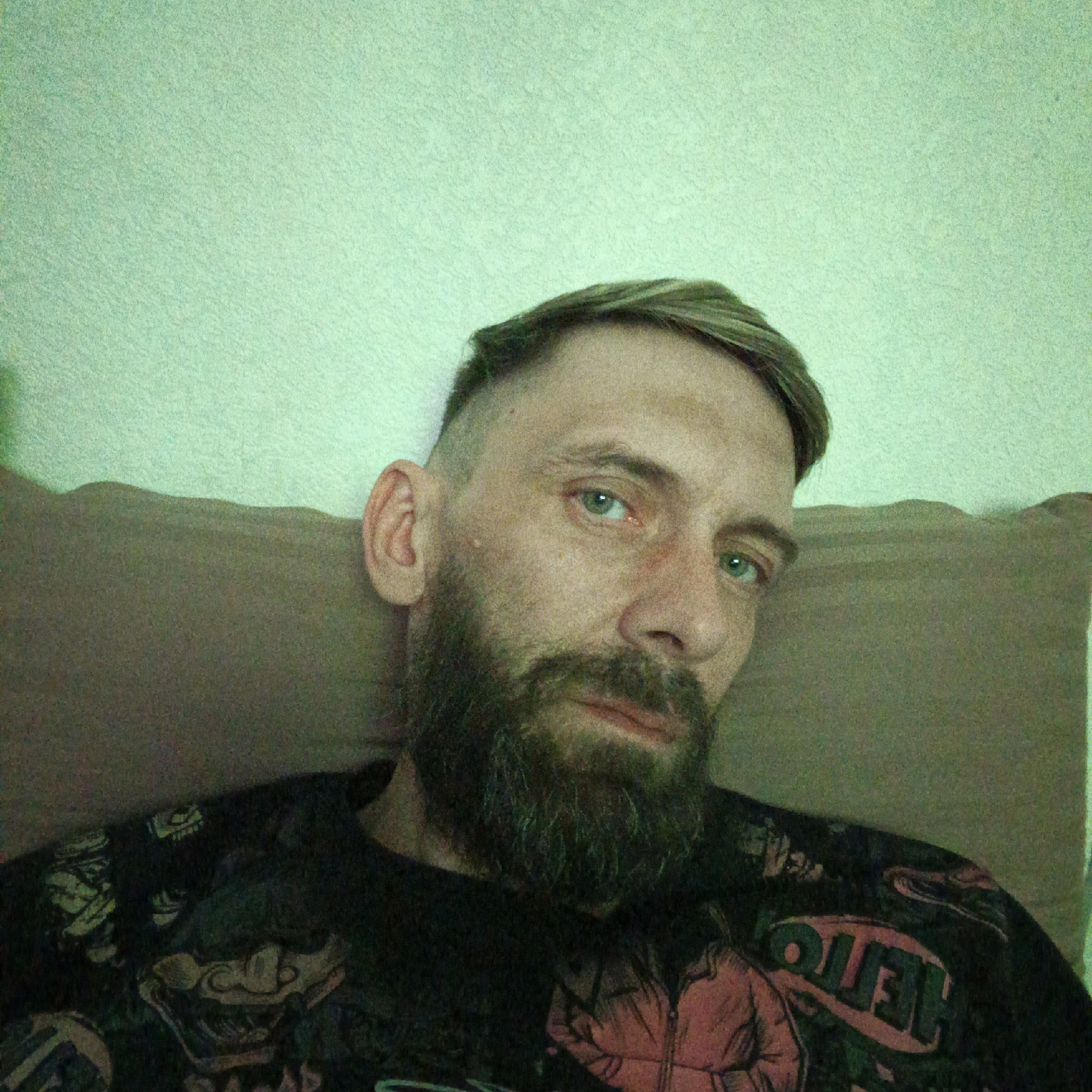 A selfie of a bearded man with short, light-brown hair lying on a brown pillow or cushion. He has pale skin, blue eyes, and a serious expression. He is wearing a dark t-shirt with colorful graphics, possibly including the word "HELLO" and cartoon-like faces. The background is a light green wall. The photo is taken indoors. The location in the city of Марковский, Russia is not discernible from the image.