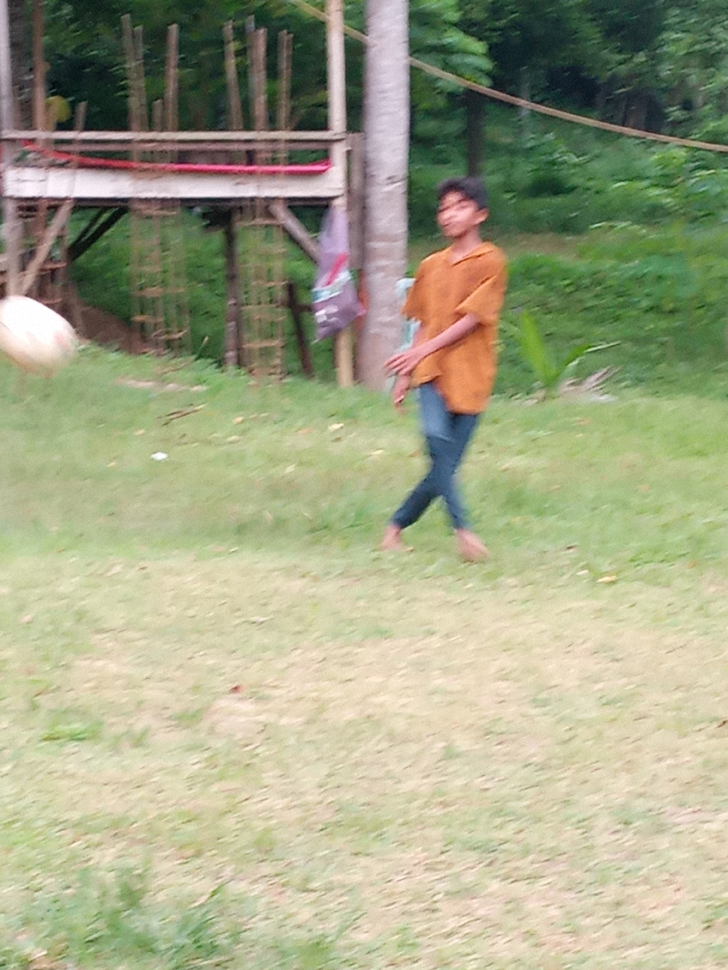 The image shows a young person, likely a boy, wearing an orange shirt and dark blue pants, walking across a grassy area. The person is captured in motion, with their left leg extended forward and their right leg bent behind them. Their head is turned slightly to their left, and their facial features are indistinct due to motion blur. In the background, there is a wooden structure, possibly a raised platform or a playground, constructed with natural wood materials. Behind the structure, dense green foliage suggests a natural, possibly wooded, environment. To the left of the frame, a pale, rounded object is visible, also blurred, which could be a ball or a similar spherical item. The overall scene appears to be an outdoor setting in a rural or semi-rural area, consistent with the environment found in Lhokseumawe, Indonesia. The lighting suggests it is daytime, with soft, diffused light.