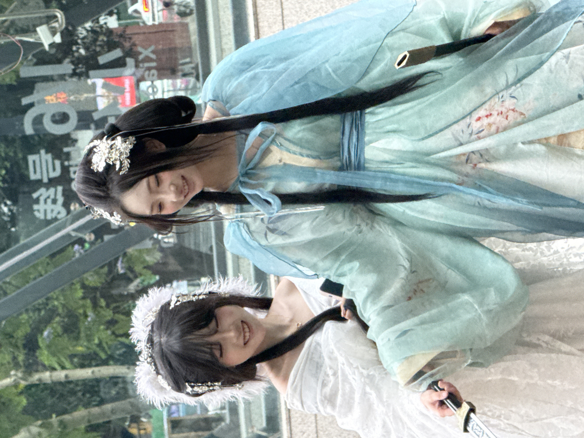 In Yuzhong District, China, two women stand closely together, one facing up and the other facing down. The woman in front wears a white dress and a feathered crown. She grasps the handle of a sword in her right hand. Behind her, the other woman wears a light blue, traditional Chinese dress and has floral ornaments in her hair. A sheathed sword is visible behind her neck. The reflection in a building behind them shows distorted images of the surrounding urban environment, including trees, building facades, and a partial sign with Chinese characters.