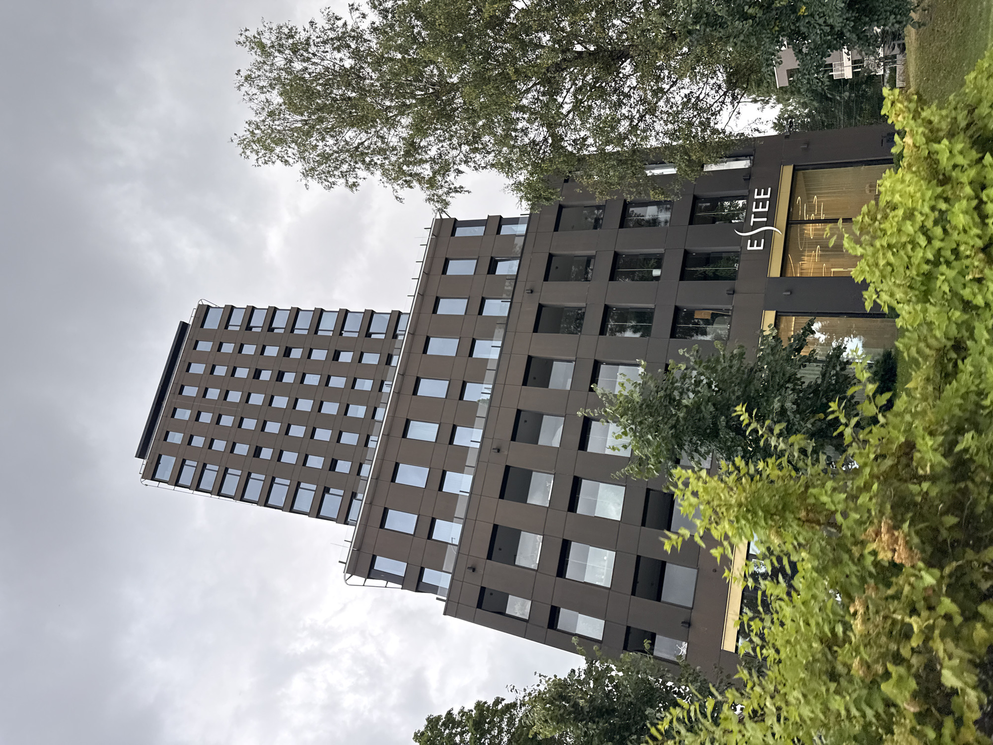 A low-angle, medium shot captures a modern, dark brown, multi-story building with a distinctly angular design, tilting upward against a gray, overcast sky. The building features numerous large, rectangular windows that reflect the sky, and on the lower floors, there are large glass storefronts with glowing circular light fixtures visible inside. A prominent sign on the lower section of the building reads "ESTEE" in white stylized lettering. Lush green trees and foliage in the foreground partially obscure the lower levels of the building, creating a contrast between the natural elements and the contemporary architecture. The scene appears to be located in Tallinn, Estonia, specifically in the Rotermann Quarter, a district known for its renovated industrial buildings and modern architecture. There are no people or vehicles visible in the image.