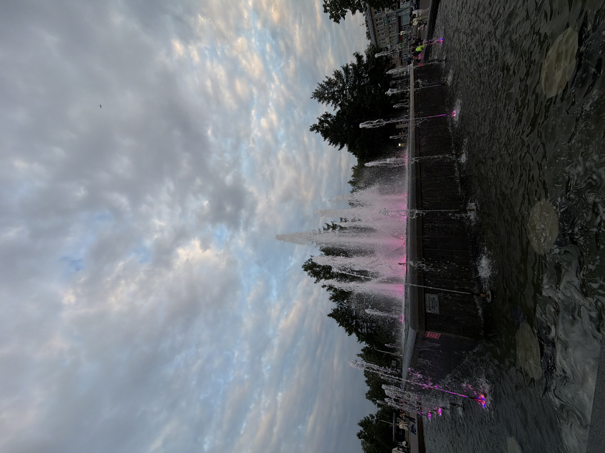 The image depicts a large, multi-tiered fountain in the city of Chaikovsky, Russia, illuminated by pink lights. The fountain is situated in a square with trees in the background and buildings visible on the right. The sky is overcast with clouds. Several people are present in the foreground and midground, some walking and others near the fountain. The water from the fountain is spraying upwards and outwards, creating a dynamic visual effect. Some of the water is also illuminated by the pink lights. The overall scene suggests an evening or late afternoon setting.