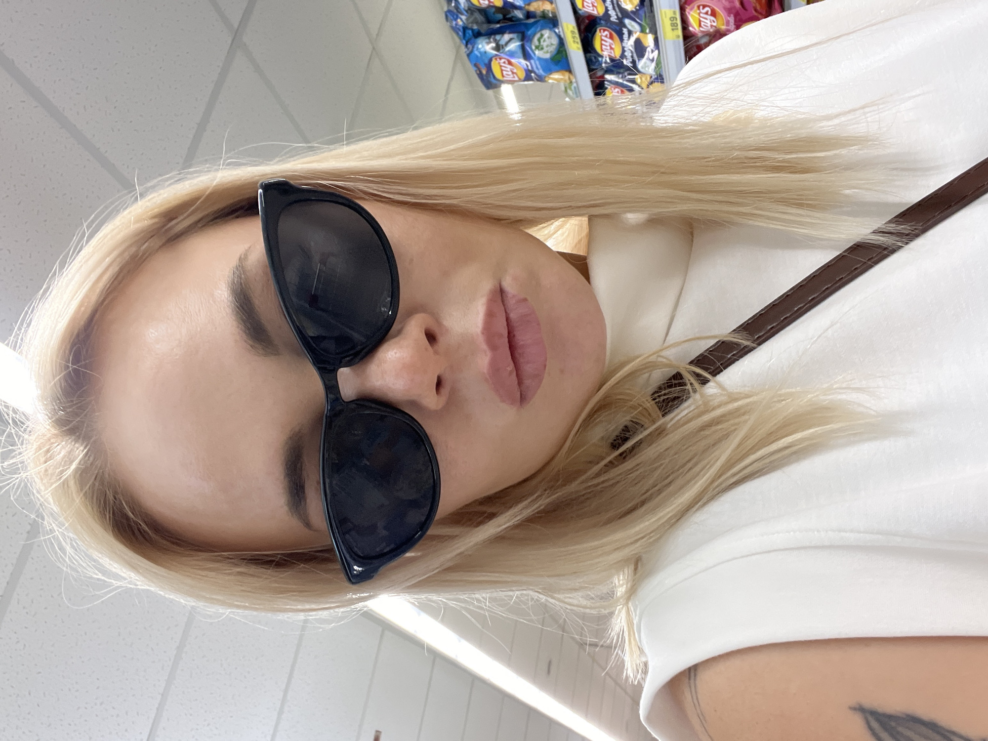 A woman with blonde hair and sunglasses is taking a selfie in a supermarket aisle. She is wearing a white sleeveless top. Behind her, shelves are stocked with bags of chips, including Lay's. The lighting is bright, suggesting an indoor environment.