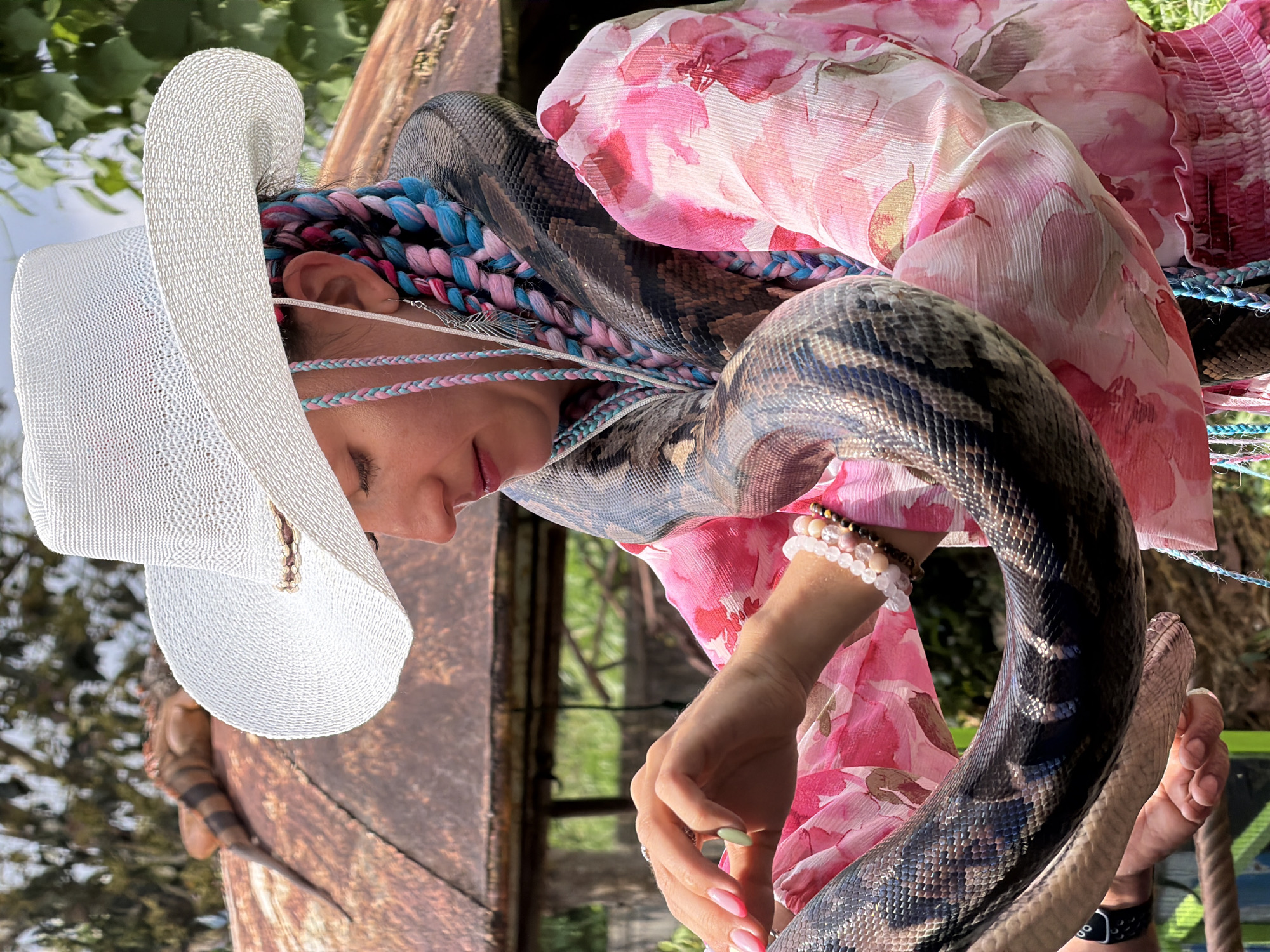 A woman wearing a white cowboy hat and a pink floral dress is holding a large python that is wrapped around her neck and arms. Her hair is styled in blue and pink braids. In the background, an iguana is visible on top of a rusty structure. The scene appears to be outdoors, with greenery and a cloudy sky. The woman is looking down, and her hands are gently touching the snake. She is wearing bracelets on her wrist, and her fingernails are painted pink and light green.