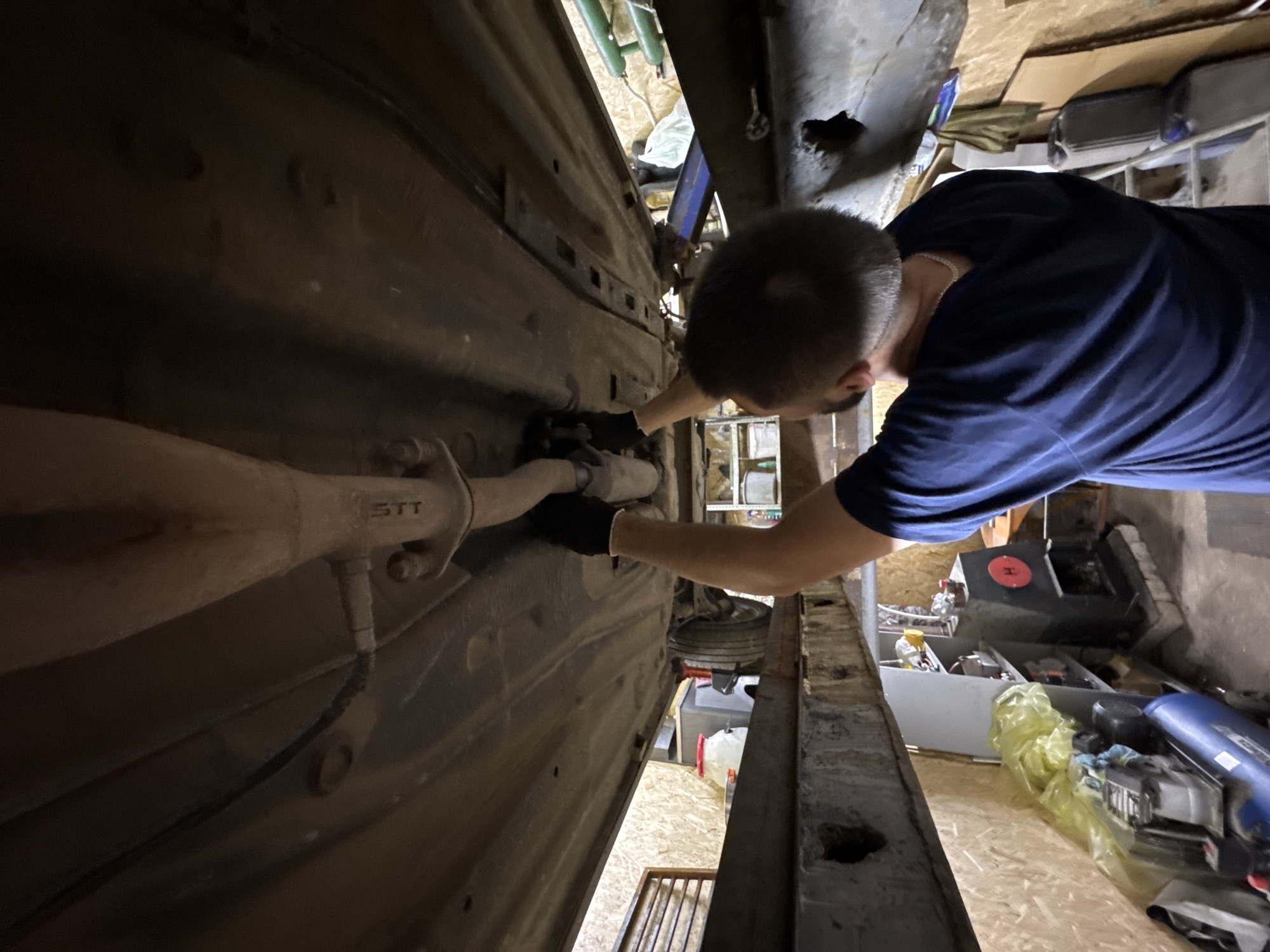 The image captures a scene within a workshop or garage, purportedly located in Perm, Russia, focusing on a male individual working beneath a vehicle elevated on a lift. The man is viewed from the back, displaying his head, shoulders, and arms. He has short, light-colored hair and a short beard. He wears a dark blue long-sleeved shirt and a silver chain around his neck. Both hands are covered with black gloves and are actively engaged with the exhaust system of the vehicle, specifically at a flange connection point on the exhaust pipe, which bears the inscription "STT" on a supporting bracket. The undercarriage of the car exhibits signs of use, including dirt and minor corrosion. A portion of a vehicle wheel is visible in the background, towards the left. The surrounding environment consists of walls paneled with light-colored wooden planks. The workspace contains various items, including a blue air compressor on the concrete floor, a yellow plastic bag, metal shelving units stocked with containers and other items, and a dark-colored stove or heater featuring a red circular emblem, situated on a raised platform. The lighting appears to be artificial, illuminating the work area beneath the car.