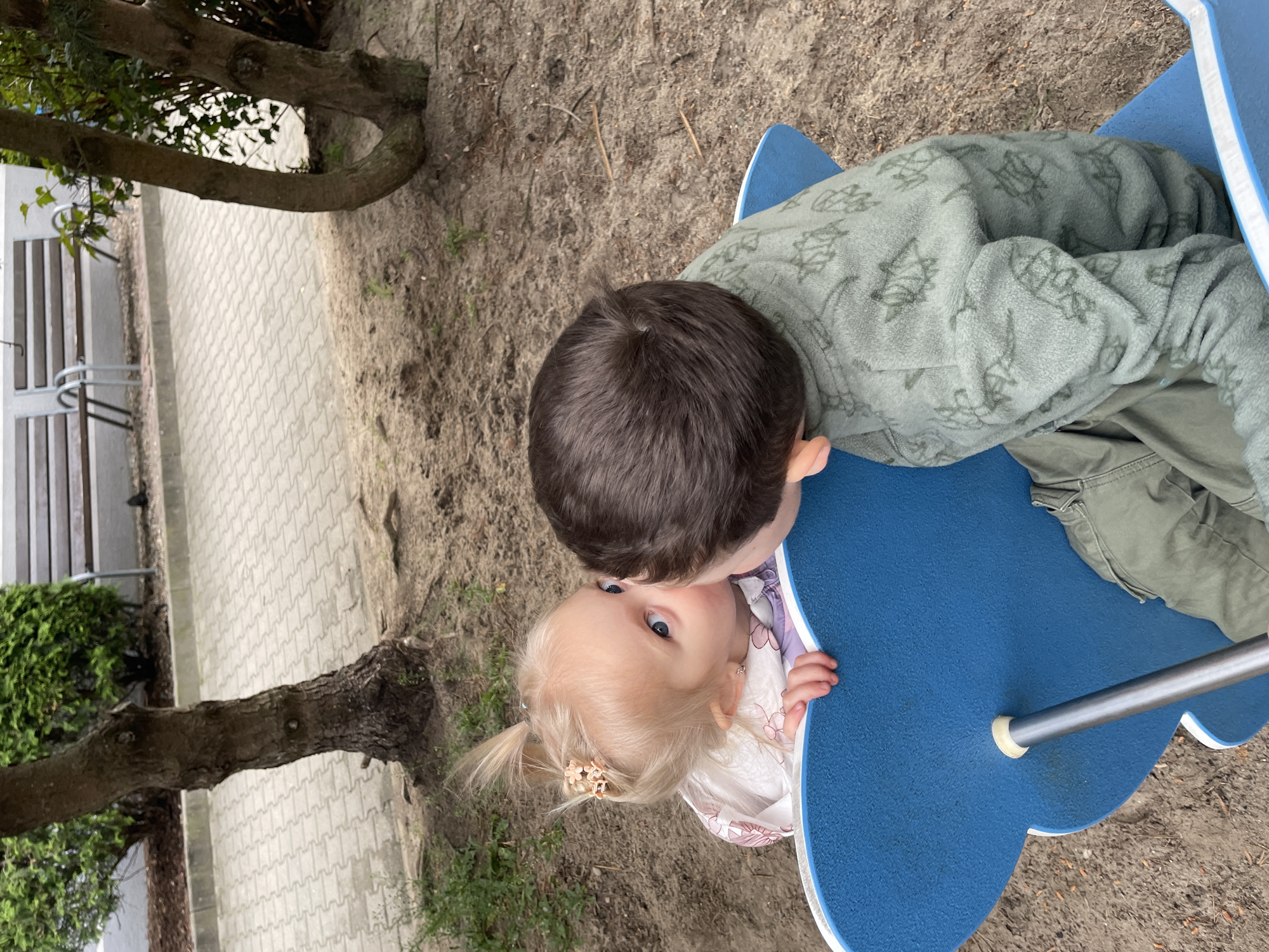 Captured from an overhead perspective, the image portrays a fair-skinned toddler with blond hair and blue eyes, seated on a blue playground seesaw. The child is adorned with a floral hair clip and a patterned white blouse. A male child, presumably older, with brown hair and clad in a green printed jacket, is seated on the opposing side of the seesaw. The surface below the seesaw appears to be dirt or sand. In the backdrop, a tree trunk is visible, alongside a paved walkway, a bench, and some greenery. The city of Unknown, Unknown is not discernible from the available visual information.