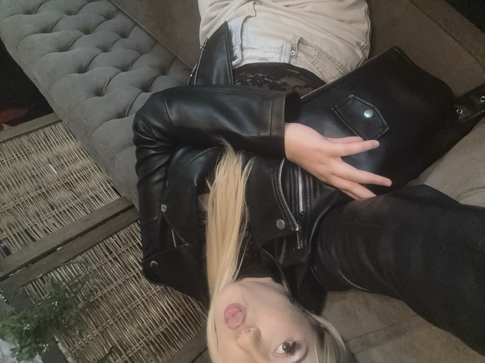 A young woman with long blonde hair is captured in a close-up, upward-angled shot, lying on what appears to be a grey tufted sofa or bench. She is looking towards the camera with a playful expression, her tongue slightly out. She has a visible nose piercing on her right nostril.
The woman is wearing a black leather jacket over a black lace top, and light-colored jeans or pants are visible around her waist. Her legs, partially seen, are covered in dark pants or leggings. Her right arm rests on a black bag, possibly a shoulder bag or clutch, which has a small flap with a silver snap button closure.
The setting includes the grey tufted upholstery of the sofa/bench she is on, and a woven straw or rattan mat is visible on the floor below and to her left. A small amount of green foliage or a plant can be seen on the mat in the lower left corner. The lighting is dim, suggesting an indoor or evening setting. The overall mood of the image is casual and relaxed. This scene is set in Stara Zagora, Bulgaria. No visible text is present in the image.
