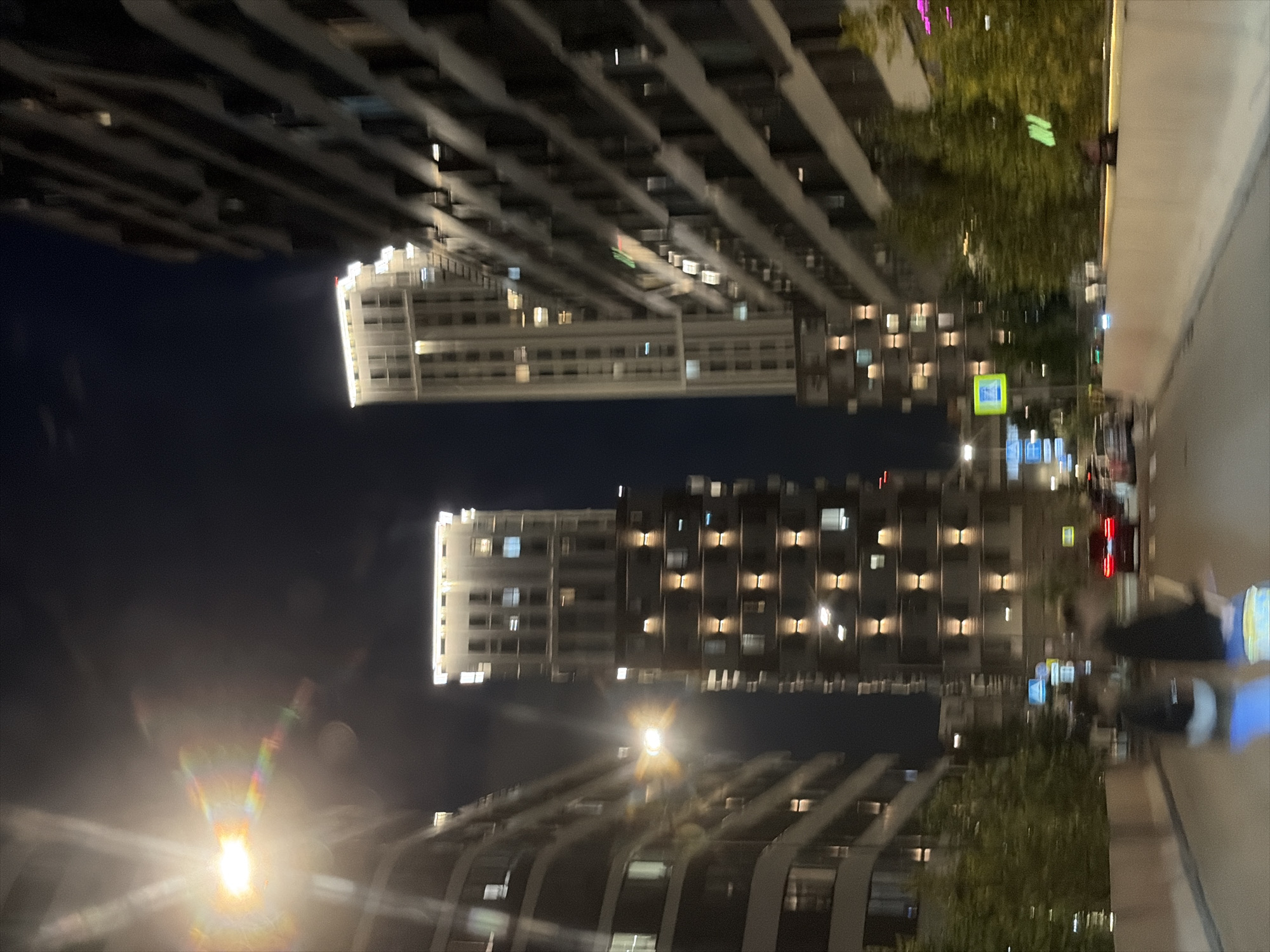 The image depicts a night scene in a modern urban environment, likely a residential or business district in Moscow, Russia. The composition is characterized by tall, contemporary buildings with illuminated windows, suggesting a well-developed infrastructure. The foreground shows a road with blurred motion, indicating that the photograph was taken from a moving vehicle. Two indistinct figures, possibly pedestrians or cyclists, are visible in the lower half of the frame, their forms blurred by movement. A red car is also present on the road, its brake lights illuminated. The presence of streetlights and building illumination creates a vibrant, albeit somewhat hazy, atmosphere due to the nighttime conditions and potential lens flare from a bright light source in the upper left quadrant of the image. The overall impression is one of a bustling city at night.