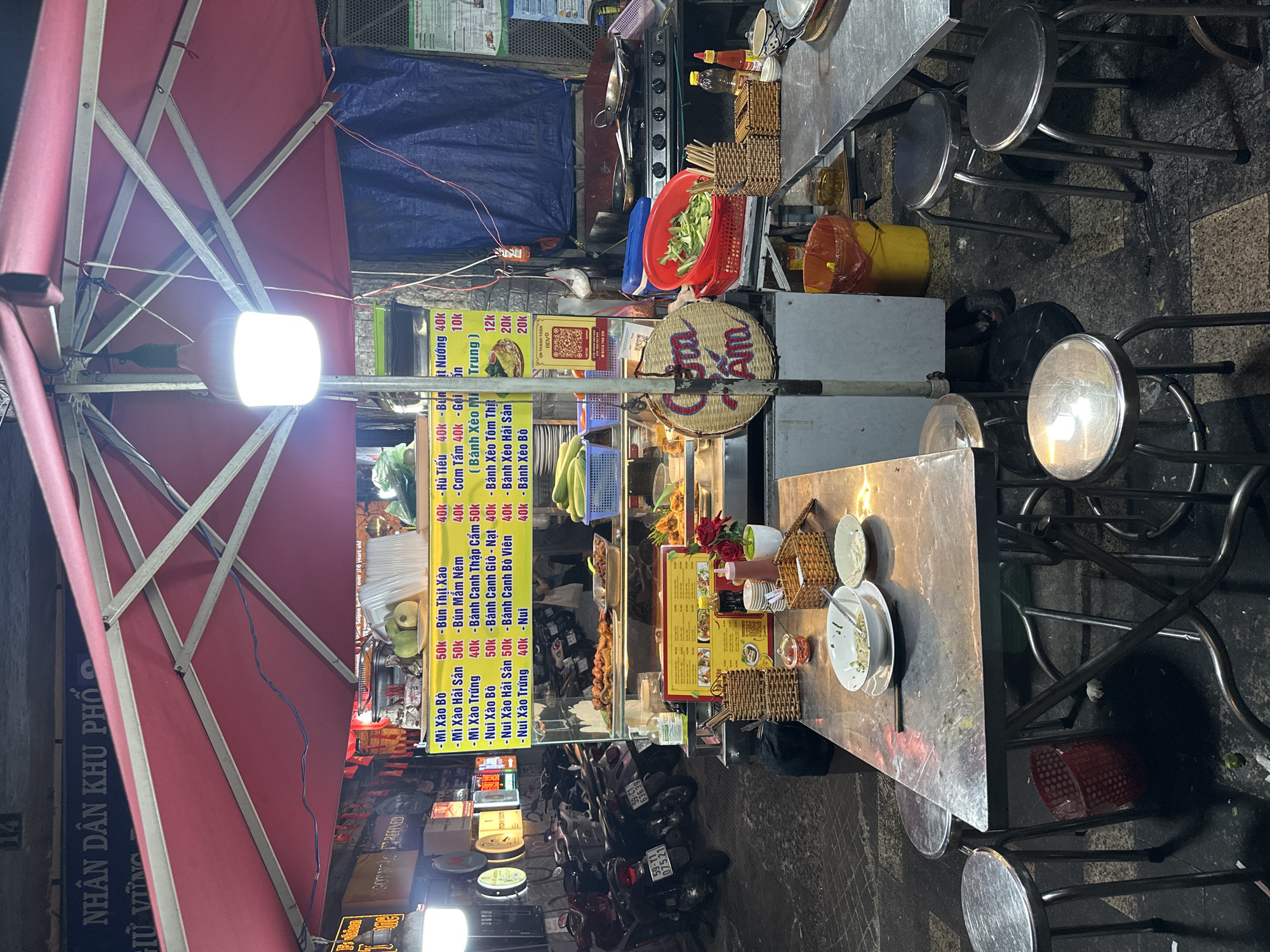 The image depicts a street food vendor in Thủ Đức, Vietnam. The vendor stall is covered by a red canopy. A bright light illuminates a menu board displaying various dishes such as "Mì Xào Bò" (stir-fried beef noodles), "Bún Thịt Xào" (stir-fried meat noodles), and "Bánh Xèo" (Vietnamese pancake), with prices listed in Vietnamese Dong. Several metal tables and stools are set up for customers. Motorcycles parked nearby suggest the scene is in a busy street. Dishes and condiments are visible on the tables.