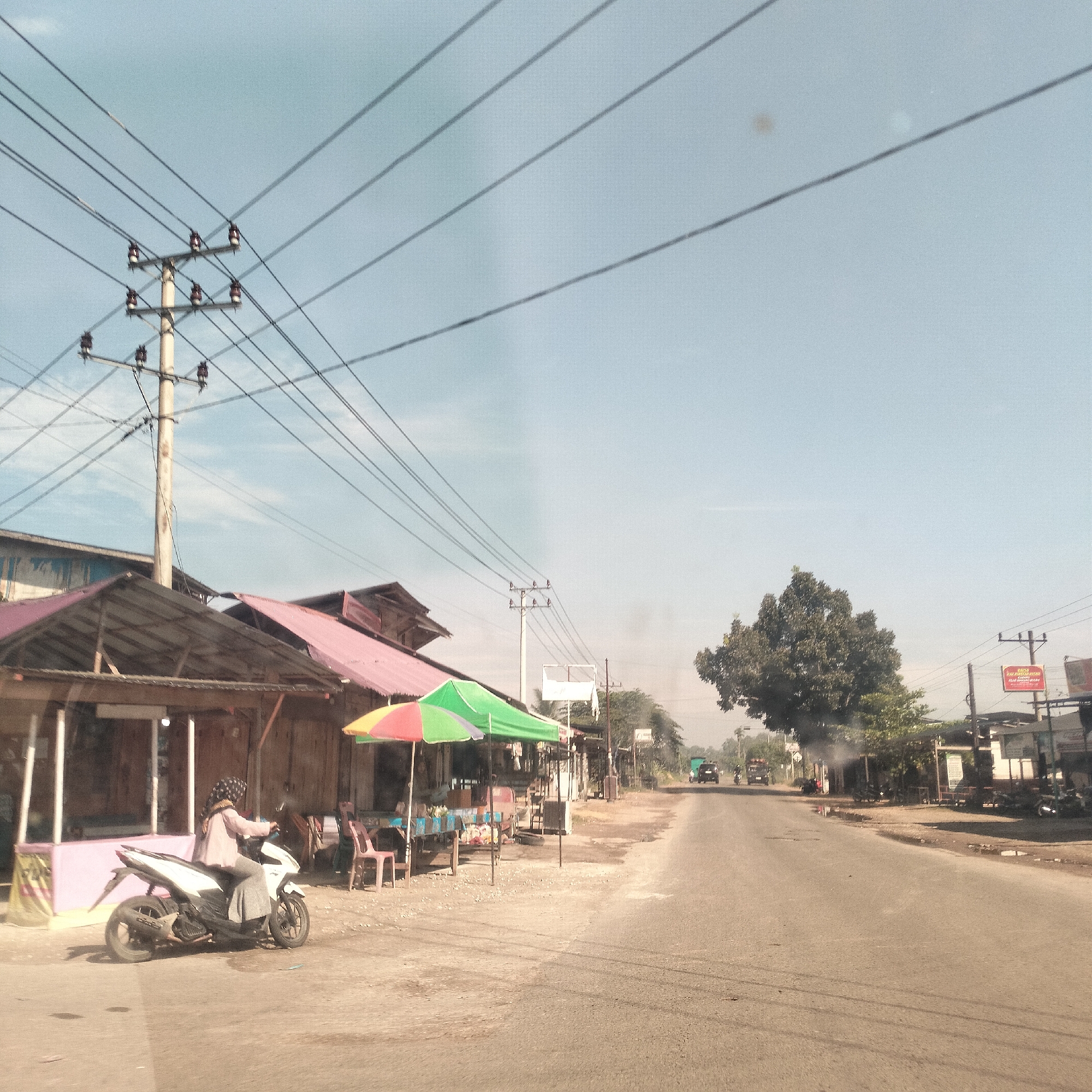 The image depicts a sunny daytime scene along a paved road in what appears to be a semi-urban or rural area, consistent with Lhokseumawe, Indonesia.
In the foreground on the left, a person wearing a light-colored top and a patterned headscarf is seated on a white scooter, facing right. The scooter is parked on a dusty, unpaved area next to simple, open-front wooden structures with light pink roofs, suggesting small shops or eateries. Adjacent to these structures are two colorful market umbrellas, one red and yellow, and another larger green and orange striped one, providing shade over an outdoor seating area with plastic tables and chairs.
Prominently featured on the left and extending across the upper portion of the sky are numerous power lines originating from a tall utility pole equipped with multiple ceramic insulators. The sky above is clear and light blue, indicating bright, fair weather.
A paved road occupies the center and right side of the frame, stretching into the distance and appearing somewhat hazy. The ground immediately beside the road is unpaved and dusty. Along the right side of the road, more basic buildings, scattered trees, and additional utility poles are visible. A large, dense green tree stands out on the right in the mid-ground, with faint smoke or haze visible around it and further down the road, possibly from a vehicle or a small fire. In the far distance, several vehicles, including trucks and motorcycles, can be seen on the road. A red sign on the right side of the road, though mostly illegible due to distance, appears to indicate a "WARUNG MAKAN" (eating stall or restaurant), further suggesting the presence of local eateries. The scene portrays typical daily activity along a Southeast Asian roadside.
