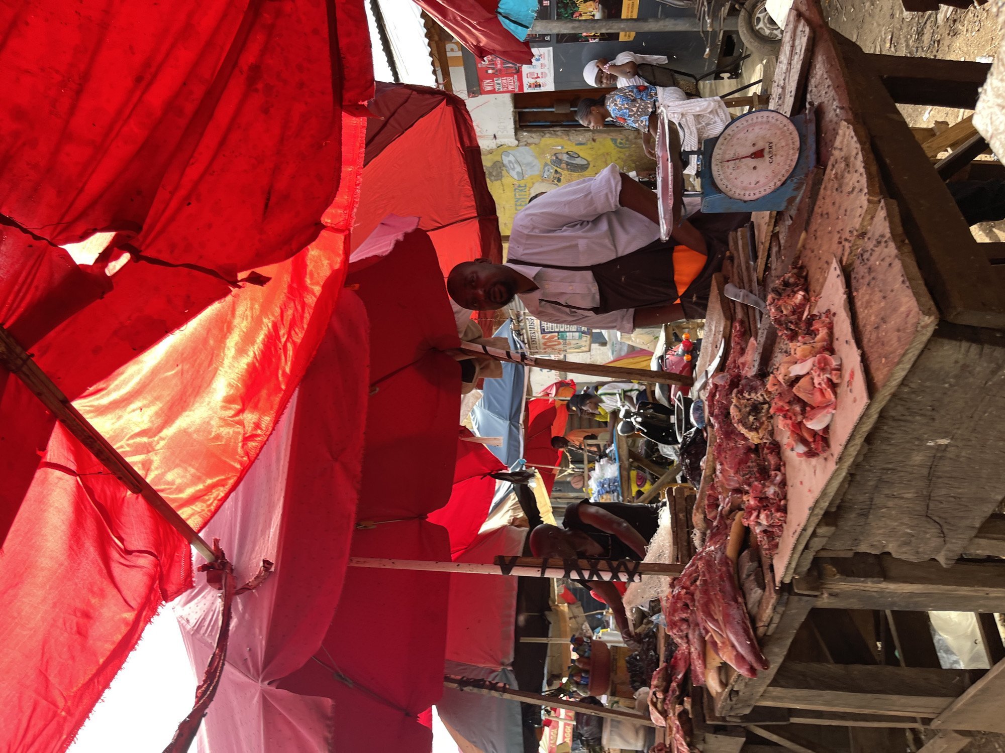 A medium shot captures an outdoor market stall in Rafin Par, Nigeria, with a focus on a butcher and his wares. The stall is covered by a bright red canopy, with sections of fabric appearing worn and slightly torn. In the foreground, a wooden table displays a substantial amount of raw meat, some of which is cut into pieces. A vintage mechanical scale, blue with a white face, rests on the table, positioned to the right of a butcher.
The butcher, a dark-skinned man wearing a striped light-colored shirt and a brown apron, stands behind the table, looking towards the camera. He is positioned slightly off-center to the right. To his right, a woman wearing a patterned blue and white dress is seated, and further to the right, another woman in a white headscarf is also seated. In the background, more market stalls and people are visible, suggesting a bustling environment. The ground appears to be dirt. Several motorcycles and a small vehicle are also present in the background, adding to the sense of a busy market day. The lighting suggests it is daytime, with sunlight filtering through the canopy.
