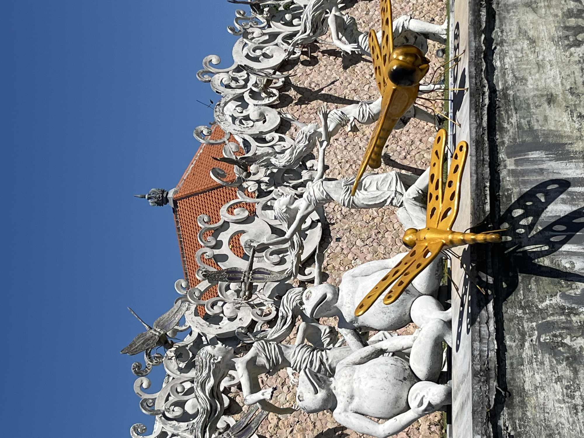 The image shows an outdoor art installation in Sukawati, Indonesia, against a bright blue sky. In the foreground, two large, golden dragonfly sculptures with perforated wings are positioned on a ledge. One dragonfly is standing, while the other lies on its side. Behind the dragonflies, a row of white, stylized sculptures depicts human figures, possibly dancers or mythical beings, with flowing hair and draped garments. These figures are integrated into an ornate, white, wavy border that runs along a stone wall. Two large, white frog-like statues are seated prominently in the lower-middle section of the frame, interacting with the human figures behind them. The background features a stone wall and a tiled red roof of a building. The overall scene is one of artistic display, likely in a public space or cultural site in Sukawati.