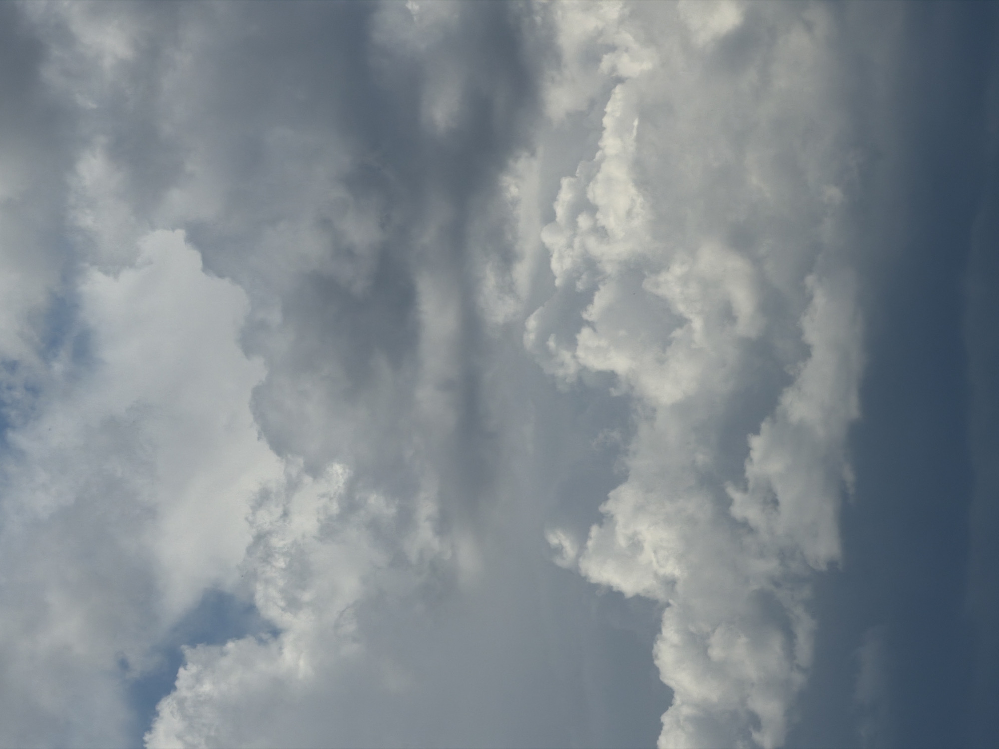 A cloudscape photograph captures a dynamic sky filled with various cloud formations. The clouds range from bright white, fluffy cumulus clouds in the lower portion of the image to darker, more diffused gray clouds higher up. Patches of blue sky are visible between the cloud layers. The lighting suggests it might be overcast or nearing sunset/sunrise. The image depicts a natural environment with no visible man-made structures or individuals. It is impossible to determine the specific location within Kolyvan, Russia, from this image alone.