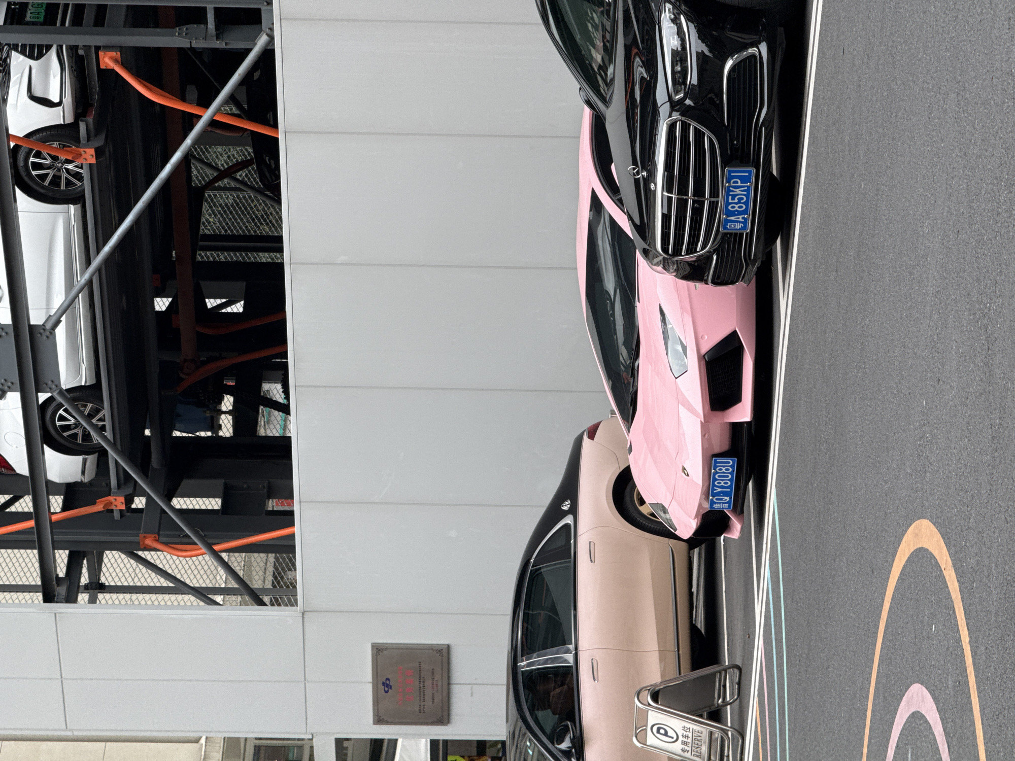 The image shows a parking area in the Liwan District, China, with several luxury cars parked. A multi-level parking structure is visible on the left, holding a white vehicle. On the ground level, there's a beige Maybach car, a pink Lamborghini, and a black car are parked. The license plate on the pink Lamborghini reads "粤Q Y808U", and the black car's license plate reads "粤A 85KPI". A parking sign with the words "专用车位 RESERVE" is visible. The parking area is marked with colorful stripes and a circle on the asphalt. A building with a plaque is in the background.