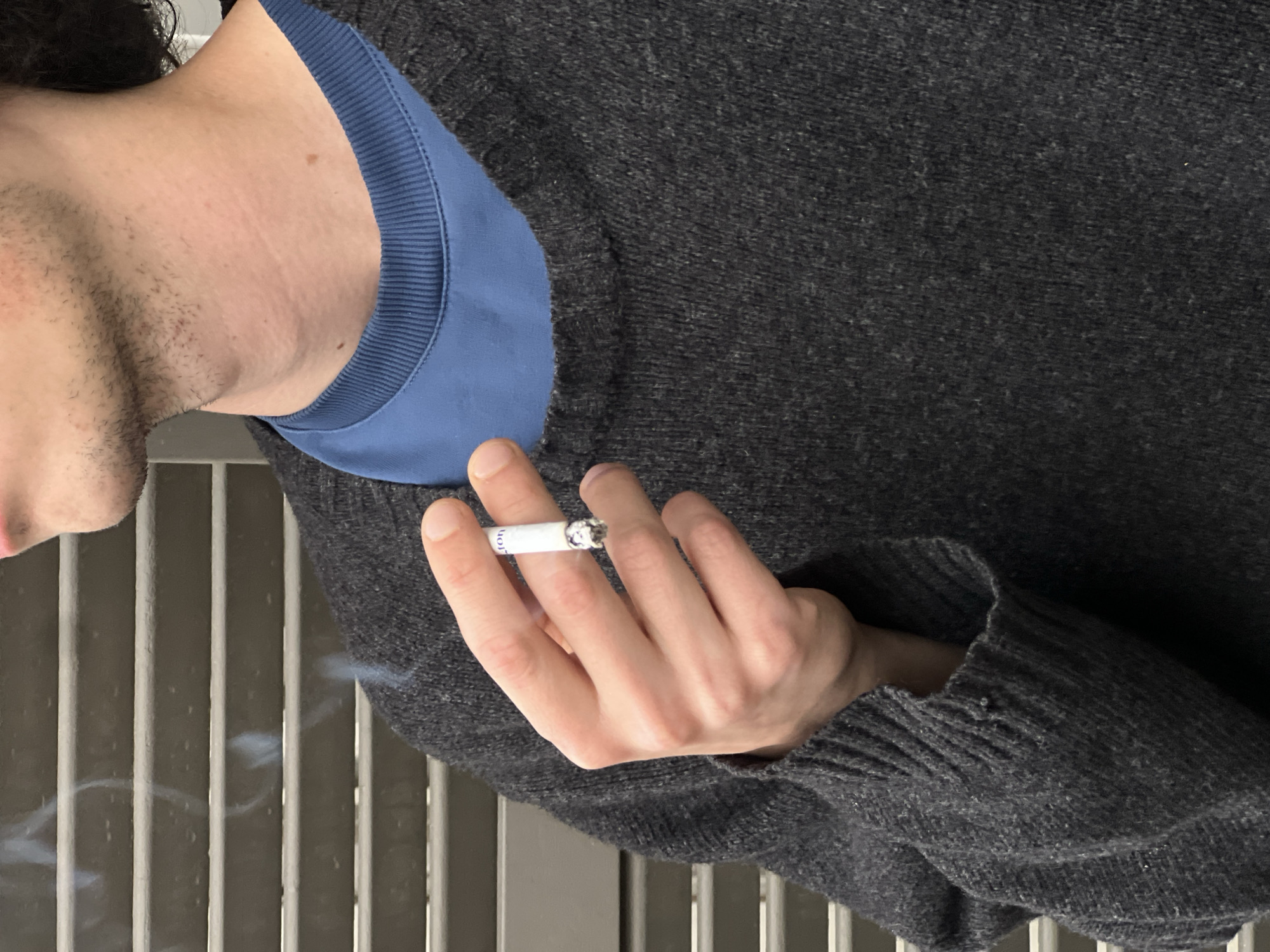 A person is holding a cigarette with smoke rising from it. The person is wearing a grey sweater over a blue shirt. The background appears to be a metal fence or railing. The person's lower face is visible, including stubble. There is no information in the image to ascertain the location in Tel-Aviv, Israel.