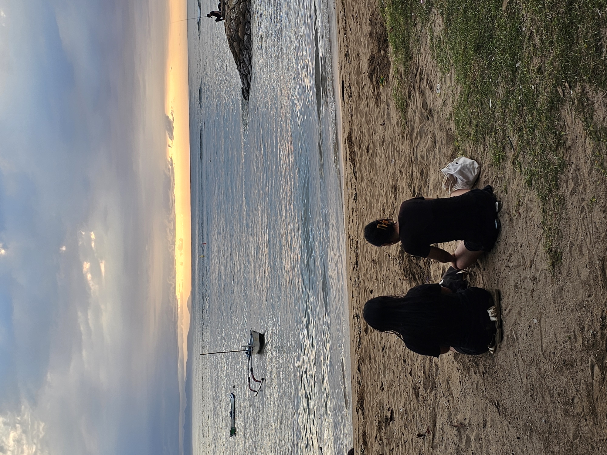 The image depicts a beach scene in Denpasar, Indonesia, featuring two individuals sitting on the sand near the shoreline. The setting sun paints the sky with hues of orange and gray, reflected in the calm water. In the sea, a small sailboat and other watercraft are visible. The beach transitions into a sandy area with patches of vegetation. One person is wearing a black shirt and shorts, while the other is also dressed in black attire. Their specific actions are not discernible, but they appear to be engaged in some activity on the beach.