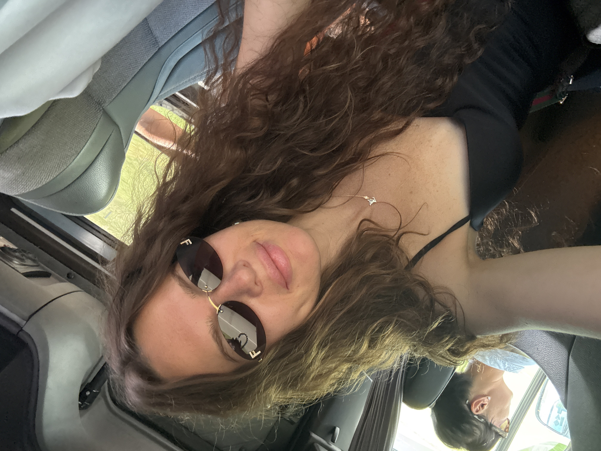 A selfie shows a young woman with long, wavy brown hair and sunglasses with gold frames, wearing a black top and a delicate necklace. She is in a vehicle, possibly a bus or van, with light gray seats visible in the foreground and background. Sunlight filters in from a window, illuminating the scene. Another person with dark hair and glasses is visible in the background, looking out the window. The location is stated to be in Onguday, Russia.