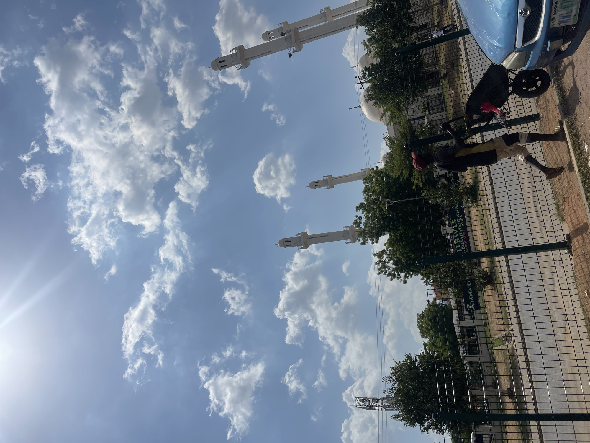 The image shows a partially obscured view of the National Mosque in Abuja, Nigeria. In the foreground, a man is visible next to a fence. He is pulling a wheelbarrow toward a blue car. The sky is blue with scattered white clouds. The mosque's minarets are visible.