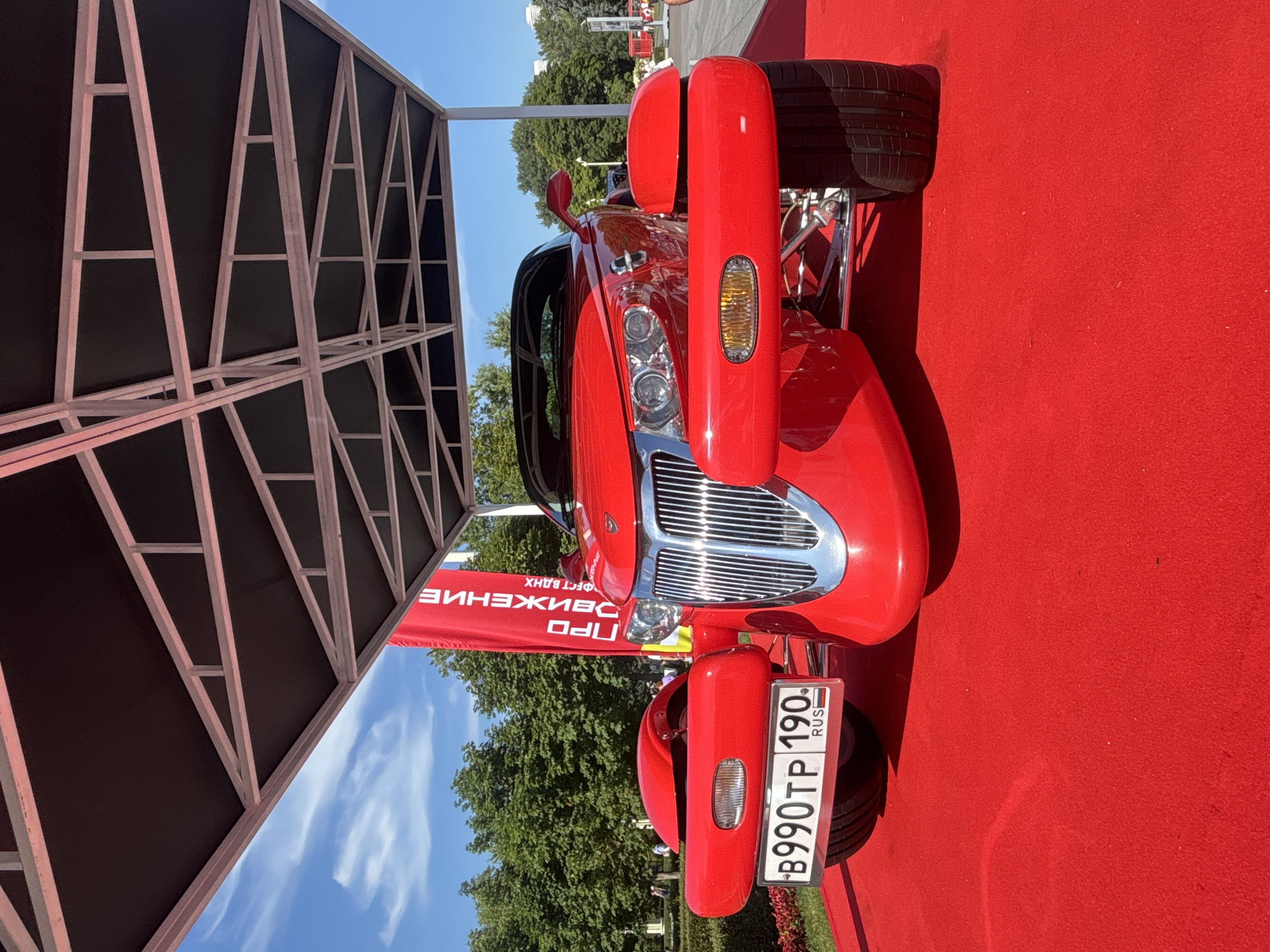 A bright red convertible car, possibly a Plymouth Prowler, is parked on a red carpet under a large, open-sided metal structure. The car features prominent chrome detailing on its grille and front bumper. The license plate reads "B990TP 190 RUS". In the background, a tall red banner with white Cyrillic text is visible. Beyond the banner, a park-like setting with lush green trees and a clear blue sky can be seen. Several people are casually walking in the background, suggesting this might be an outdoor event or exhibition in Moscow, Russia. The lighting indicates it is daytime.