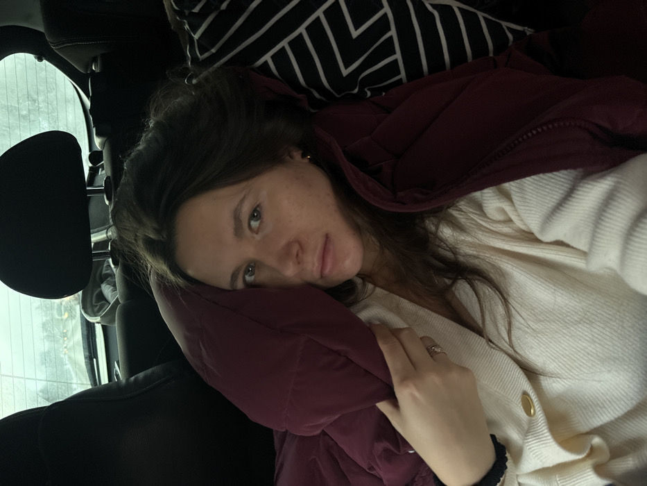 A young adult female is depicted lying or reclined in the back seat of a vehicle, reportedly in Рассудово, Russia. She has long, dark brown hair and light-colored eyes, looking directly towards the camera with a neutral expression. Her complexion is light, showing some minor skin variations. She is wearing a cream-colored, V-neck, ribbed knit cardigan, with a single gold-colored button visible on the left side of her chest. Her left hand is visible, partially holding a maroon-colored puffer garment, which appears to be a vest or jacket, and a silver-toned ring with a central clear stone is present on her left ring finger. A small gold stud earring is visible in her right ear. To her right, a black and white geometrically patterned textile, possibly a pillow or blanket, is partially visible. The dark interior of the car is visible around her, including black headrests and portions of the rear seating. Through the rear window, a blurred outdoor scene suggests a road or similar environment under overcast conditions, with distant foliage discernible.