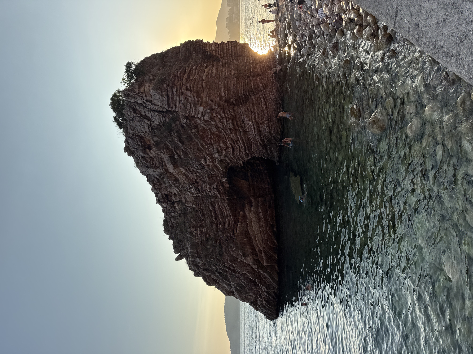 A large, layered rock formation with a natural archway stands in the sea near the shore in Kamenovo, Montenegro. The sun is setting behind the rock, casting a warm glow and creating a bright reflection on the water. Several people are visible in the shallow water near the rock, some swimming and one with a snorkel. Others are sitting or standing on the rocky shore to the right of the formation. The water is clear, allowing the seabed and rocks beneath the surface to be seen. The sky is a pale blue with a gradient towards yellow near the horizon, indicating dusk. The rock itself is reddish-brown and appears to be composed of many sedimentary layers. Small trees and vegetation grow on the top of the rock. In the distance, a hazy coastline with buildings and hills can be seen.