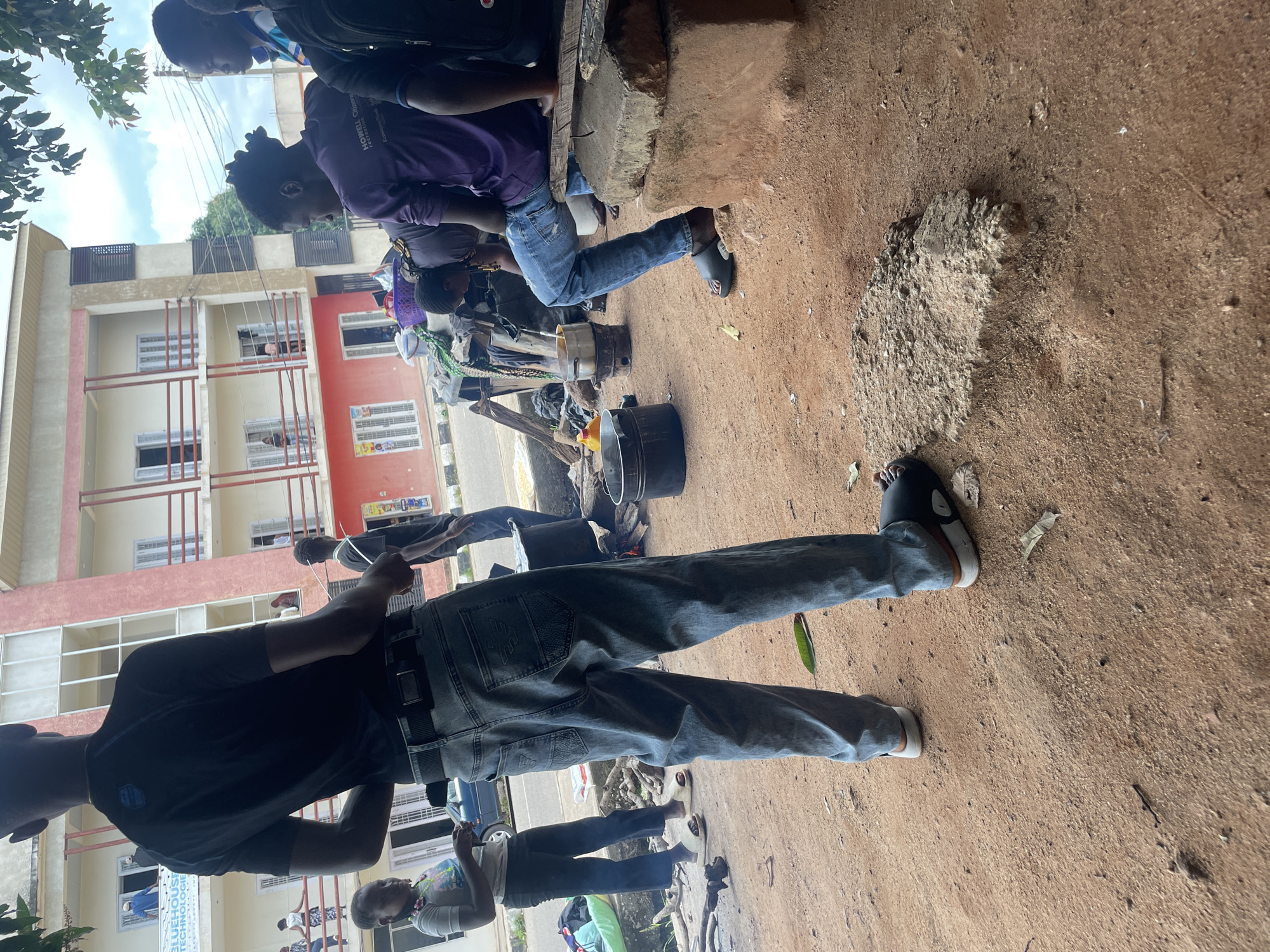 In Jos, Nigeria, a street scene unfolds with several individuals engaged in various activities around cooking fires. In the foreground, a person stands facing away from the camera, wearing a black t-shirt and blue jeans. To their left, a young woman in a grey t-shirt and dark pants is looking down, holding something in her hands. Behind them, a person in a purple t-shirt and blue jeans is seated on a raised platform made of concrete blocks. A collection of pots, bowls, and cooking utensils are arranged on the ground, some of which are placed over small fires. The environment appears to be a dusty, outdoor setting with buildings in the background, one of which has multiple balconies. The overall impression is of a communal outdoor cooking area on a city street.