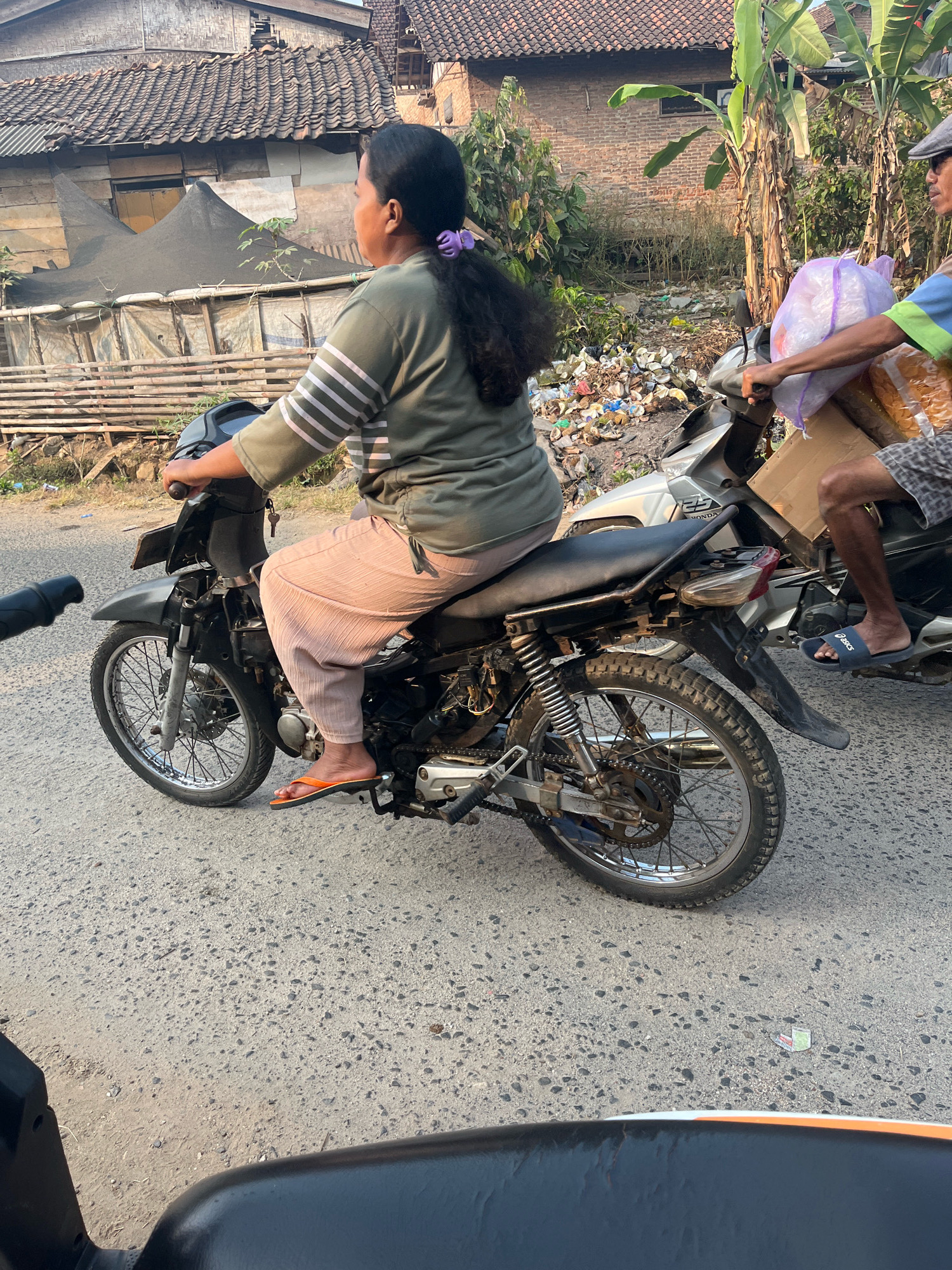 In Negeri Agung, Indonesia, a woman rides a motorcycle with her dark hair pulled back. She wears a green and white striped shirt and light-colored pants, with orange flip-flops. She is looking to her right. A second motorcycle, ridden by a man wearing a grey cap, a blue and green shirt, and plaid shorts, is to her right, carrying a large purple bag and a cardboard box. The scene is set on a paved road with a backdrop of rural homes and piles of trash, suggesting a potentially unsanitary environment.