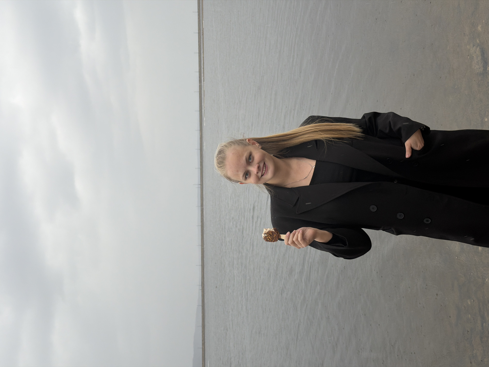 A young woman with blonde hair, wearing a black jacket, stands in an outdoor area. She is holding a popsicle in her right hand. The background consists of a flat body of water that meets a gray sky. There are visible posts lining the water. Based on the location, this image may have been taken near the shores of the Votkinsk Reservoir in Chaikovsky, Russia.