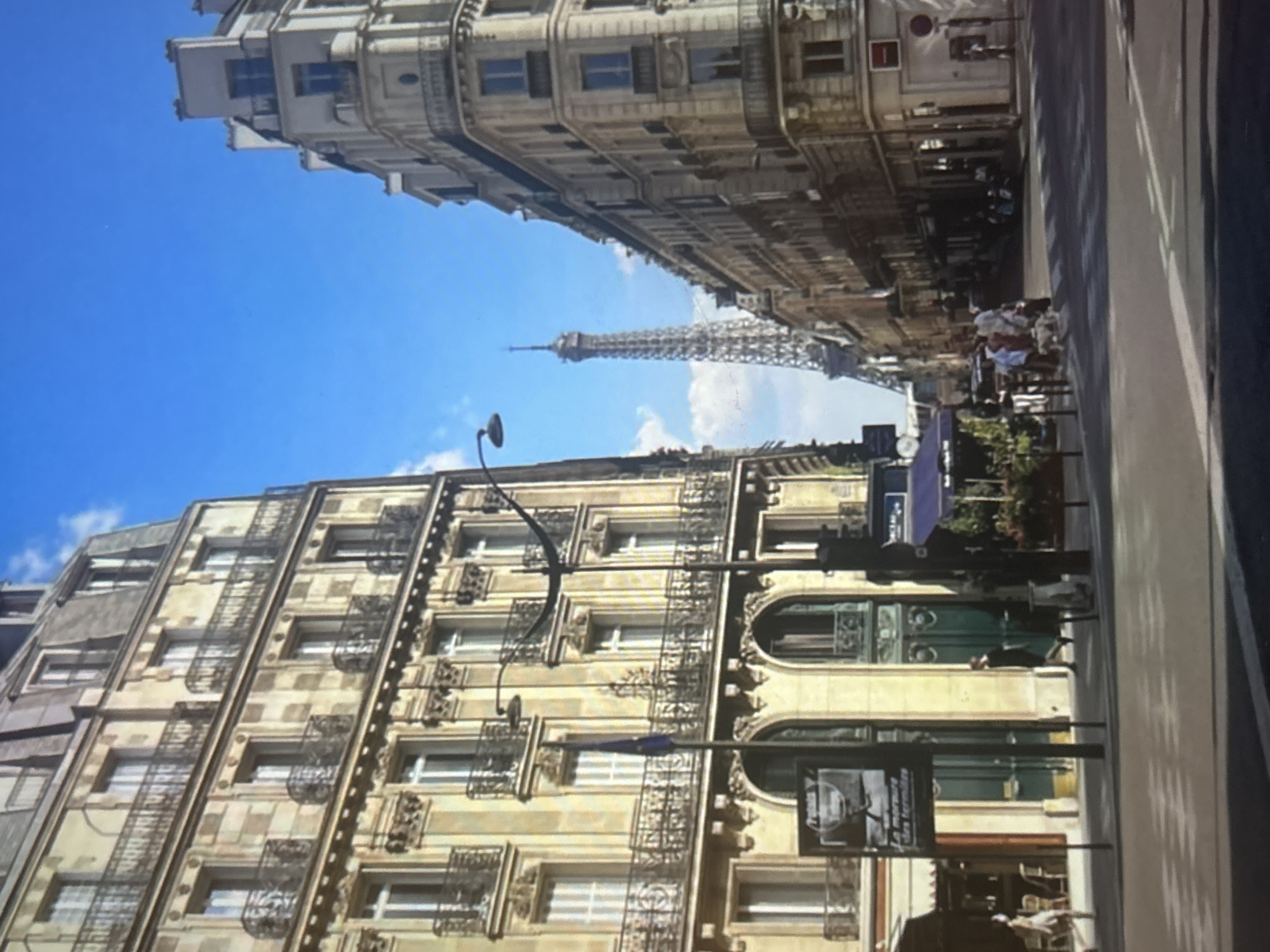 The image captures a view of a street in Paris, France, with the Eiffel Tower visible in the distance. On the left, a tall, ornate building with many windows and balconies lines the street. A flagpole with a blue flag stands in front of this building. On the right, another multi-story building with a rounded corner and balconies also fronts the street. The street itself is paved and appears to be a crosswalk area with white markings. Several people are walking on the sidewalk and crossing the street. Some are dressed casually, and there is a dog being walked. There are shops and cafes with outdoor seating visible along the street. The sky is blue with a few white clouds. The overall impression is of a sunny day in a bustling urban environment.