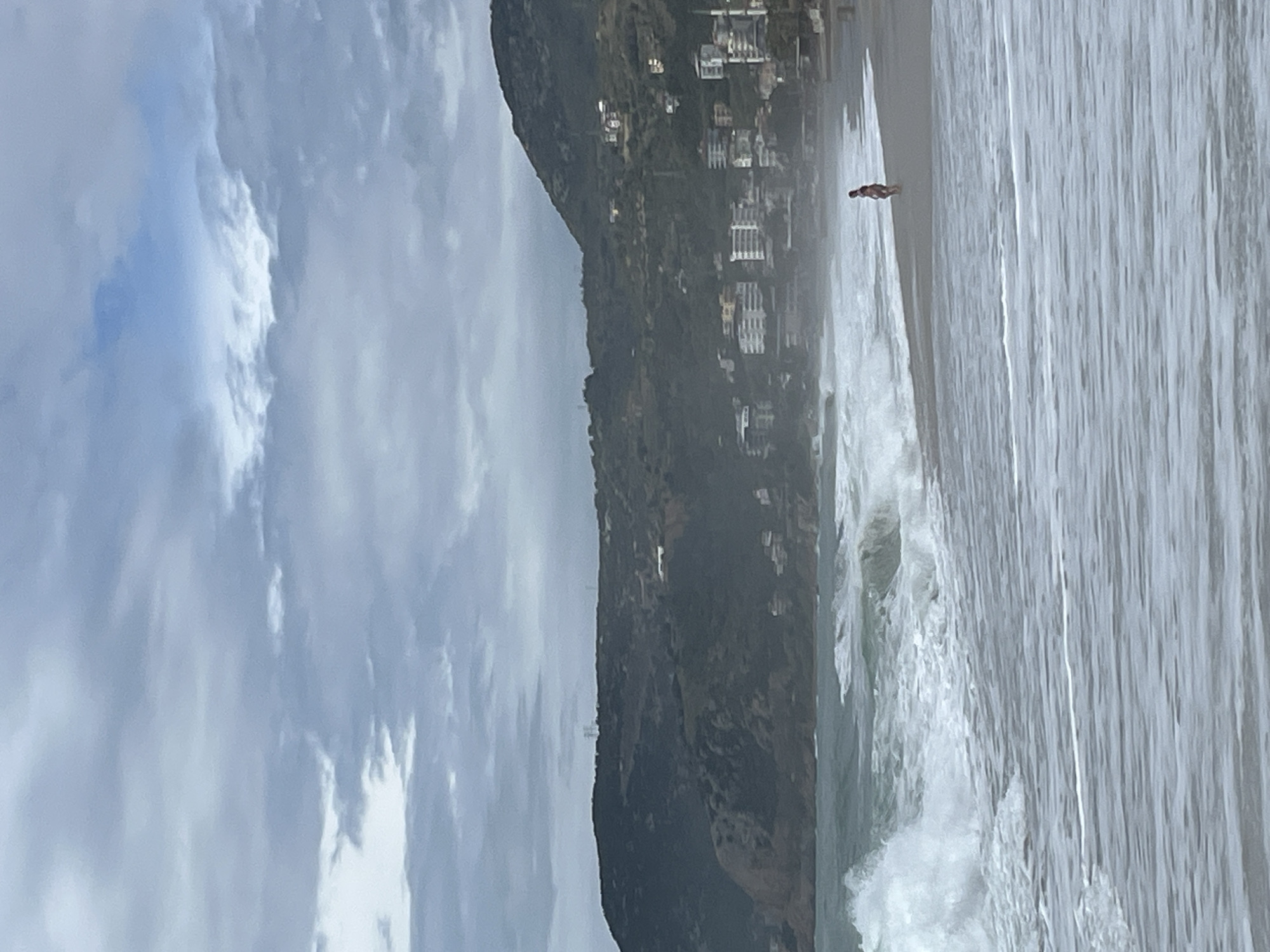The image depicts a beach scene in Alanya, Turkey. There is a mountain range adjacent to the beach that is covered in trees and has a few buildings scattered along its slope. The beach has a single person walking in the distance, while waves are crashing onto the shore. The sky is overcast with many clouds, creating a dim ambient light.