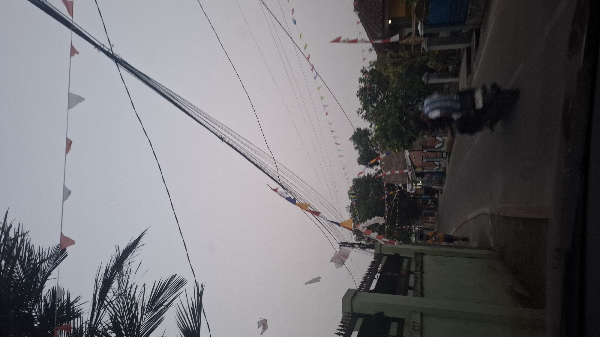 The image depicts a street scene in Ciharashas, Indonesia, under an overcast sky. Decorative flags, primarily in red and white, are strung across power lines, suggesting a celebration or national event. A paved road runs through the center of the image, with a sidewalk to the left. On the sidewalk, a lone individual wearing a yellow shirt and dark shorts walks away from the camera. A motorcycle with two riders is traveling down the road, with the rider in front wearing a blue and white striped shirt. Houses with pitched roofs line the right side of the road. Palm fronds are visible in the upper left corner. The overall impression is a street decorated for a festive occasion in a residential area.