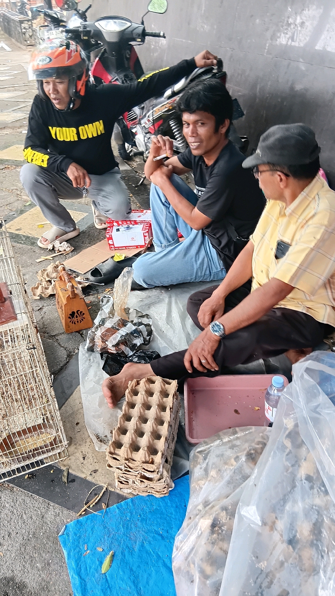 Three men are sitting on the ground near a motorcycle on a paved surface. The man on the left wears a black long-sleeved shirt with yellow text that reads "YOUR OWN" and an orange helmet. He is crouching and holding keys. The man in the middle wears a black t-shirt and blue jeans and is smoking a cigarette. The man on the right wears a yellow plaid shirt and a black baseball cap. He is wearing a watch on his left wrist.
Scattered around the men are various objects, including a white cage with birds inside, an orange wooden carved box, an egg carton, a red box, a pair of black sandals, a pink tray, plastic bags filled with what appear to be animal feed, and a bottle of water. The background features a grey wall. The scene appears to be on a street in the city of Baringin, Indonesia.