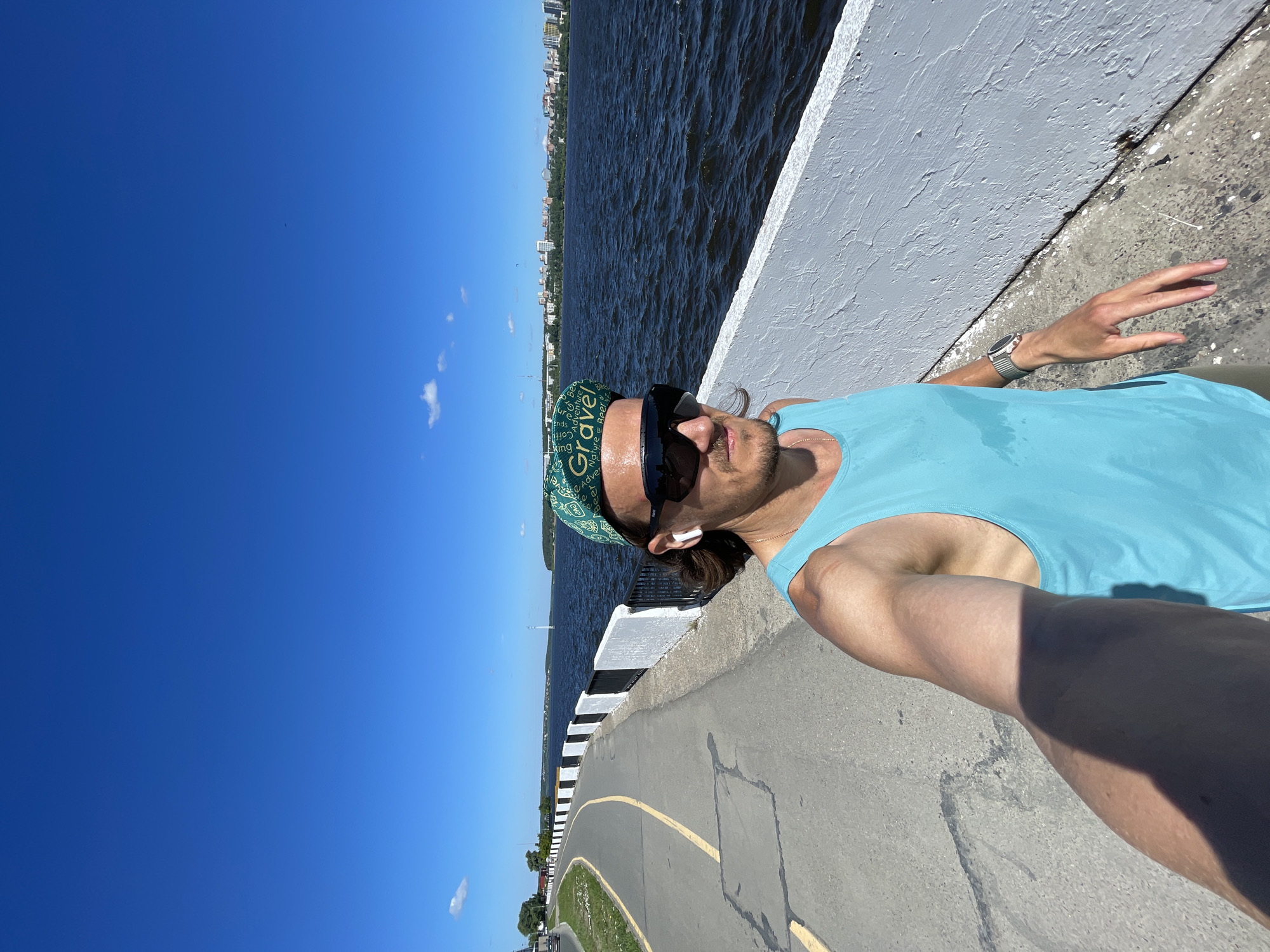 An outdoor, eye-level, medium-full shot shows a person taking a selfie while walking or jogging along a paved pathway beside a body of water. The person is wearing a teal tank top, tan shorts, sunglasses, white earbuds, and a green cap with the word "Gravel" in white lettering. The person's right arm is extended, holding the camera, and their left arm is bent with their hand relaxed at their side. A smartwatch is visible on their left wrist. The pathway has yellow lane markings and a black and white striped railing separates it from the water. In the background, a cityscape with tall buildings is visible across the water under a clear blue sky with scattered clouds. The scene appears to be in a city park or along a waterfront promenade.