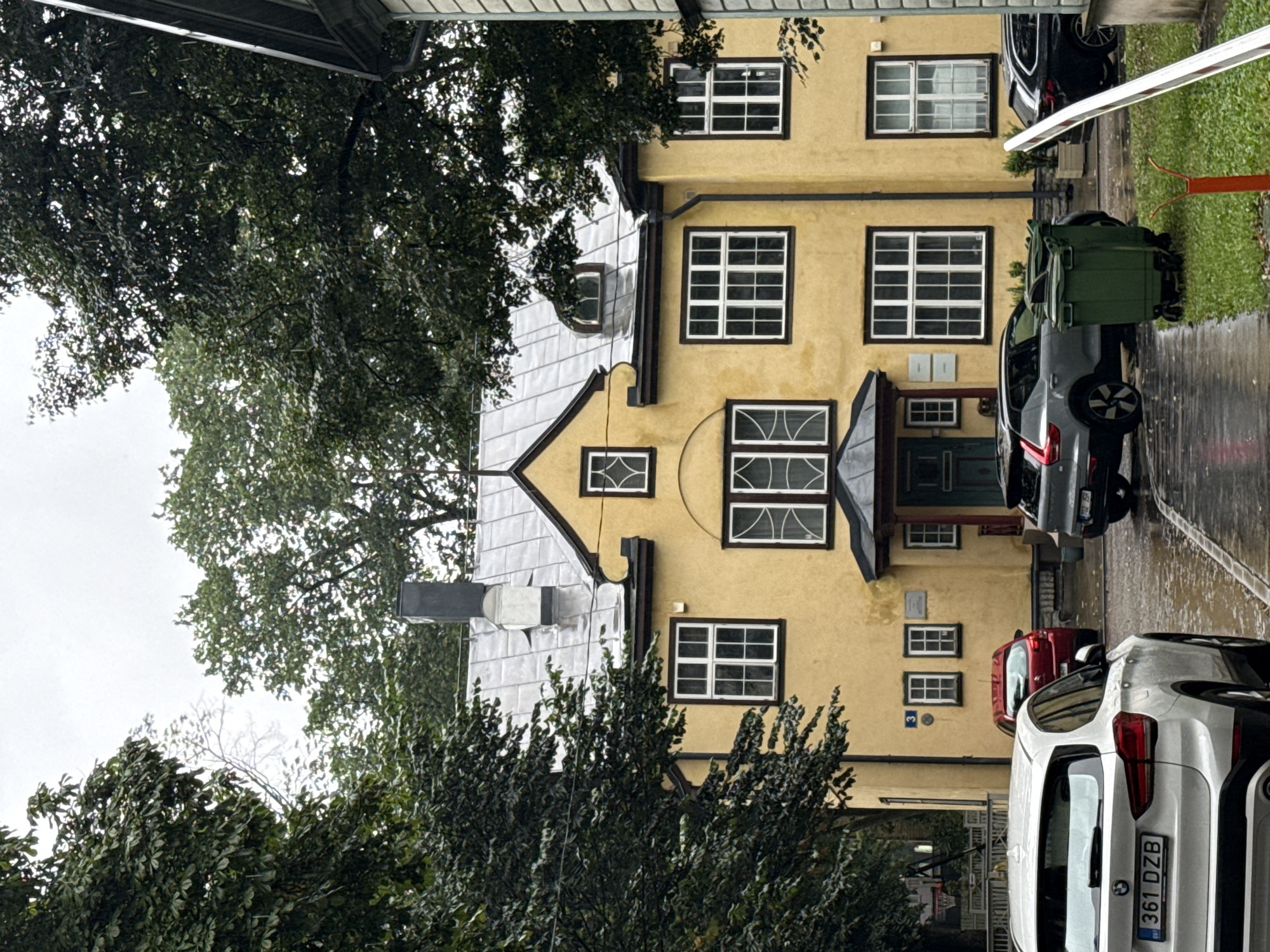In Tallinn, Estonia, a bright yellow, two-story building with a dark grey, tiled roof stands prominently. The building has numerous white-framed windows, some with decorative cross-hatching in the upper panes. A dark chimney protrudes from the roof, and a small, arched dormer window is visible on the upper roofline. Green trees with dense foliage partially obscure the upper portions of the building. In front of the building, a paved area, wet from rain, reflects the surroundings. Several vehicles are parked, including a white SUV in the foreground with the license plate "361 DZB". A red hatchback is parked further back, and a grey SUV is to the right of it. Two large green trash bins are situated near the sidewalk. To the right of the scene, a white and red striped barrier pole is visible. The overall atmosphere is overcast, suggesting a cloudy or rainy day.