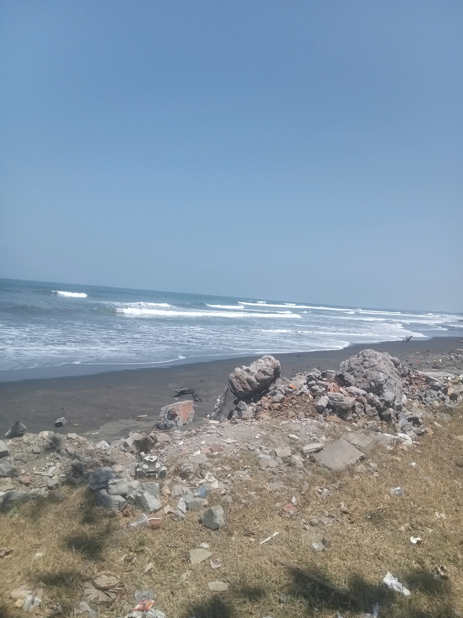 The image depicts a beach scene with a dark sand shore and the ocean in the background under a clear blue sky. In the foreground, there is a large pile of rubble and debris, composed of rocks, broken bricks, and other construction waste. Some of this debris is scattered on dry, yellowed grass. The ocean has white waves breaking on the shore. The overall impression is one of a neglected or damaged coastal area. There are no people visible in the image.
