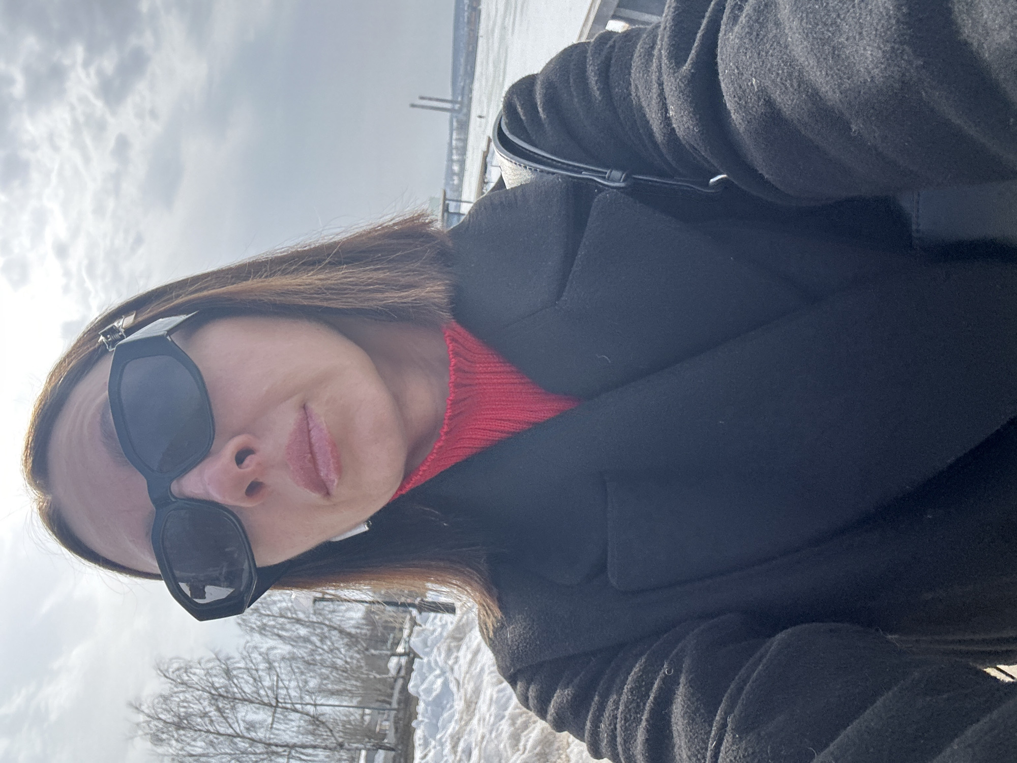A woman is depicted in a close-up selfie, looking directly into the camera. She wears large, black hexagonal sunglasses, which obscure her eyes and upper face. Her lips are slightly parted and appear glossy. She has medium-length brown hair. She is dressed in a bright red turtleneck sweater layered under a black wool-blend coat, the lapels of which are visible. A white wireless earbud is present in her left ear, and a black bag strap is visible over her right shoulder.
The background on the left shows accumulated snow on the ground and several bare deciduous trees against a bright, partly cloudy sky. To the right, a large expanse of frozen water, consistent with the Votkinsk Pond (Votkinsk Reservoir), extends into the distance. Beyond the frozen surface, industrial structures featuring two tall, slender smokestacks are visible, indicative of the Votkinsk Machine Building Plant. A metal railing, likely part of an embankment or promenade, runs along the foreground edge of the frozen water. The scene is located on an embankment overlooking the Votkinsk Pond in the city of Votkinsk, Russia, during a winter or early spring season.