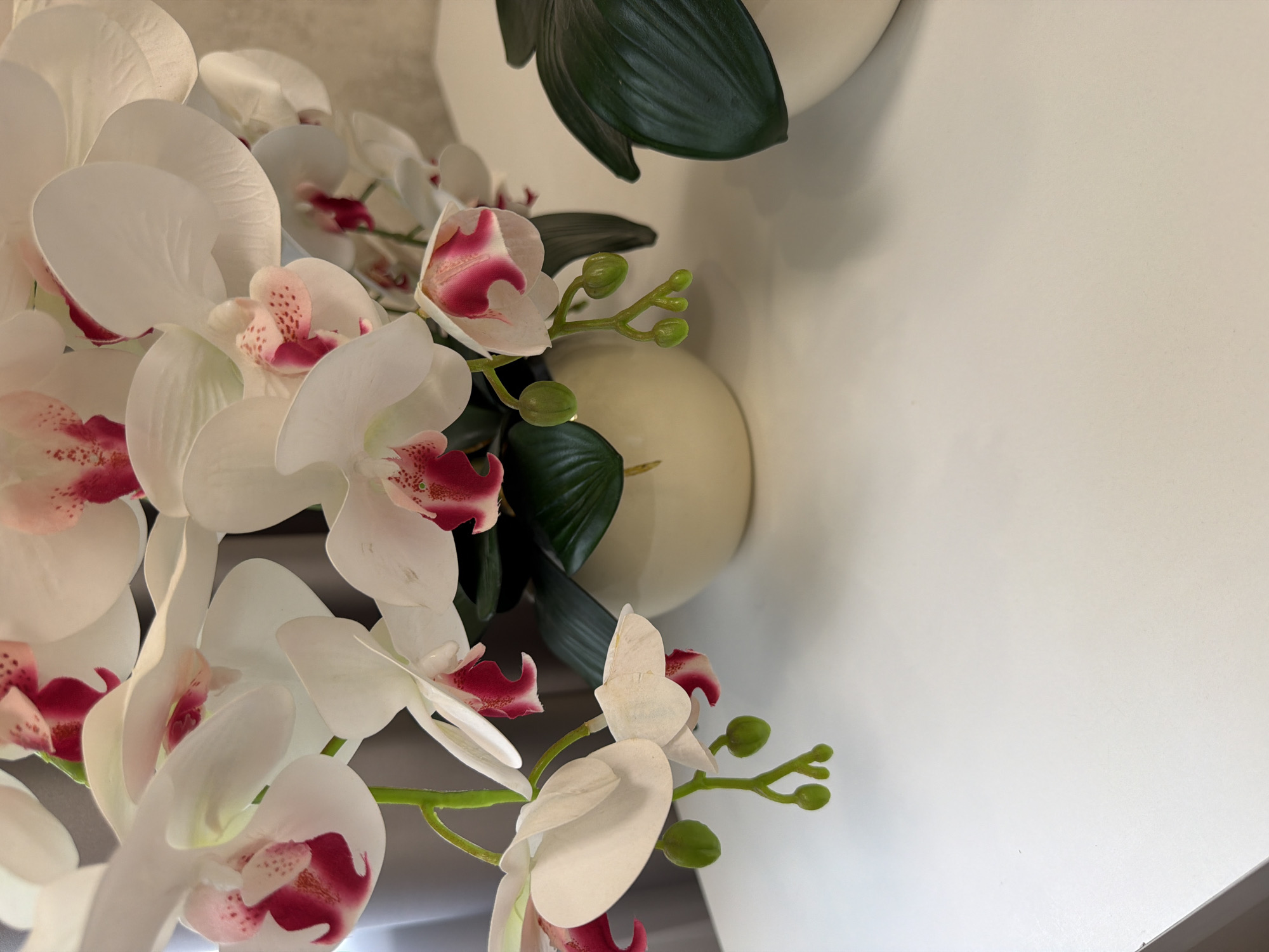 A close-up, eye-level, vertical shot features a bouquet of white phalaenopsis orchids with fuchsia centers. The orchids are arranged in a cream-colored ceramic vase, with dark green leaves visible. The bouquet is set against a blurred background of grey curtains and a wall. To the right of the vase, another cream-colored vase with dark green leaves is partially visible. The foreground shows a plain white surface, likely a table, with a shadow cast by the arrangement. The lighting is soft and even, highlighting the delicate details of the orchid petals. The image is composed to draw attention to the floral arrangement, with the white surface occupying the lower half of the frame.