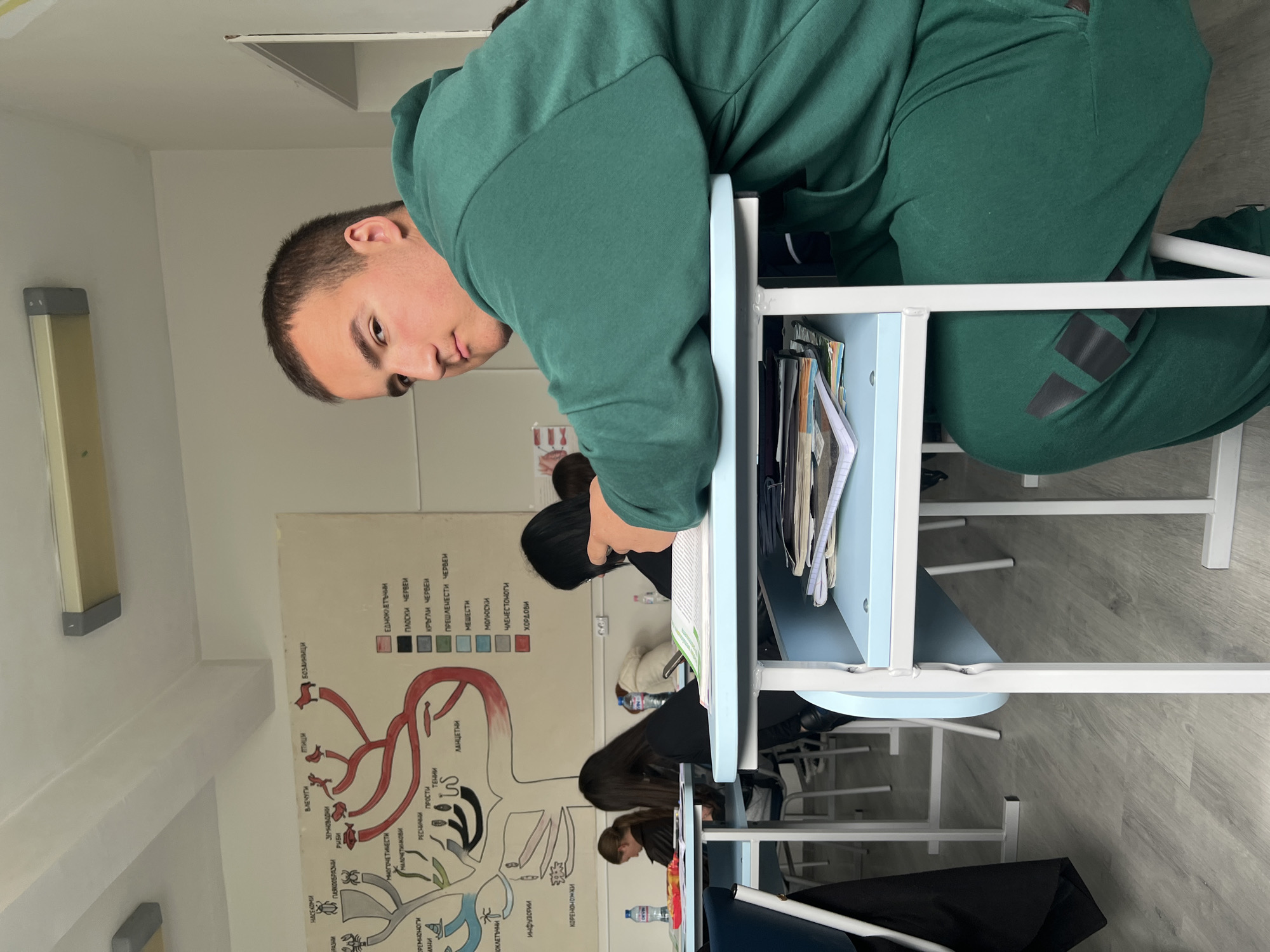 The image shows a young man in a green tracksuit sitting at a school desk in what appears to be a classroom. The desk is light blue and contains books and notebooks. In the background, there are other students at their desks. A large poster depicting biological diagrams is affixed to the wall. The OCR data suggests the poster contains text in Bulgarian, including classifications of animals. Without additional information, it is impossible to confirm the location in Krumovgrad, Bulgaria.