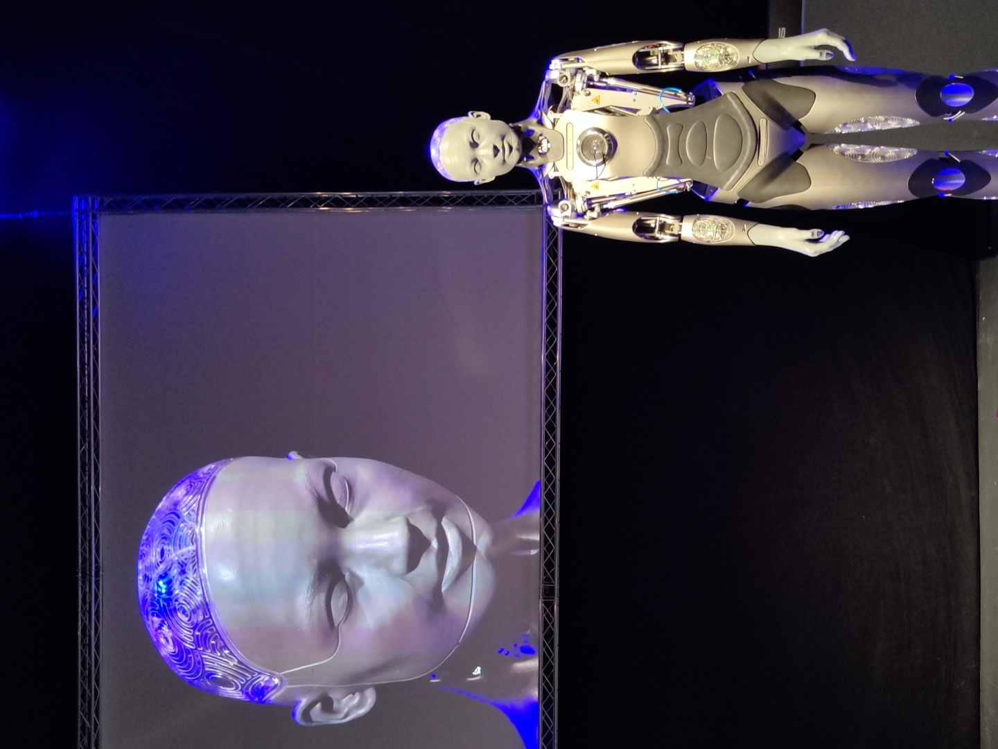 The image shows a humanoid robot standing in front of a large screen displaying a close-up of a robot's head with intricate patterns on its scalp. The robot in the foreground is silver and grey, with exposed metallic joints and blue lighting accents on its legs. The background is dark, with a black curtain or backdrop and a silver truss structure above the screen. The scene appears to be an indoor event or presentation. The image does not provide any information about the city of Haifa, Israel, or any specific location within it.